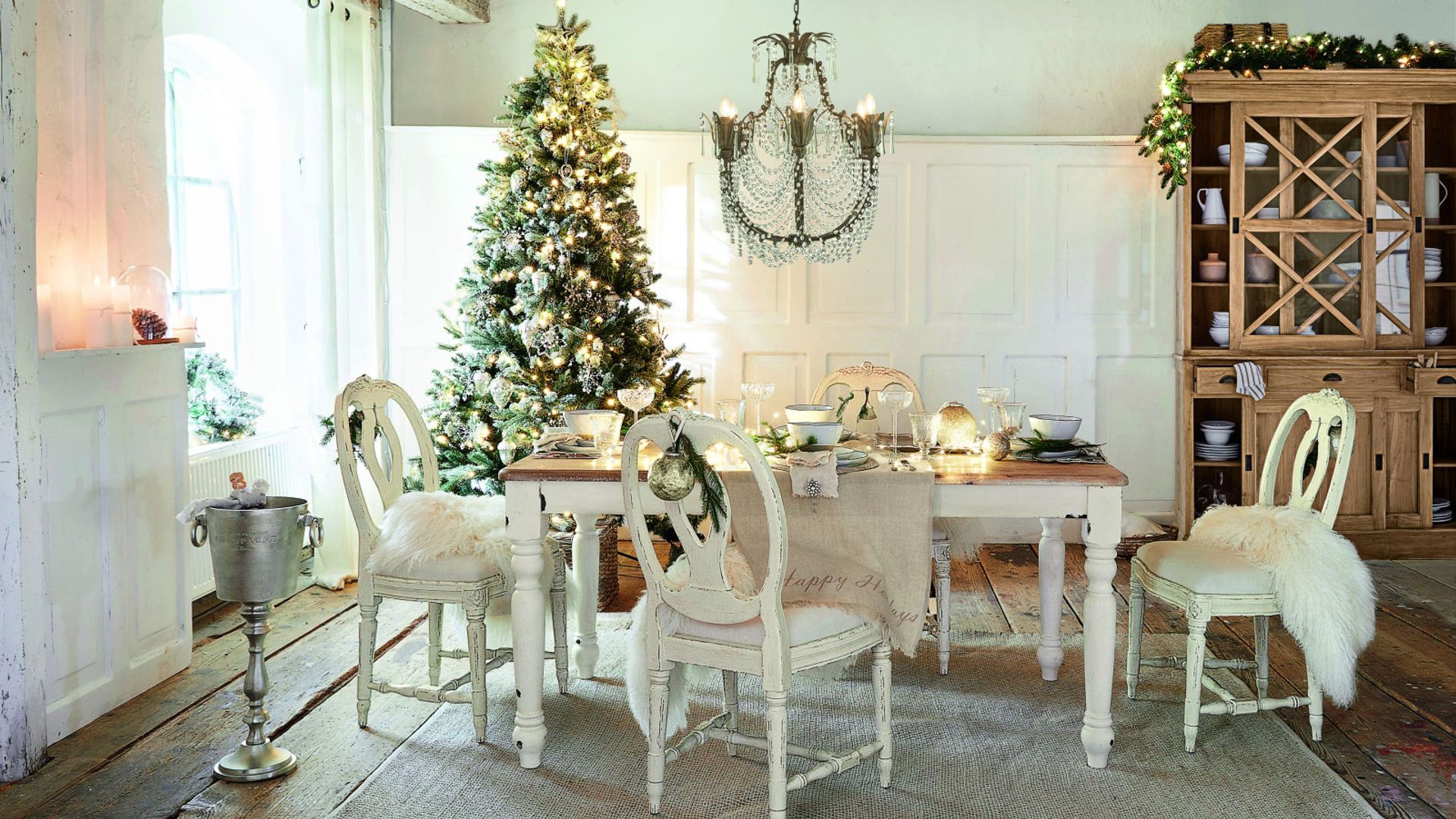 Comedor de Navidad con árbol de Navia, suelo de madera, alfombra, mesa de madera blanca con el sobre de madera, sillas blancas, con manta de pelo, lámpara de techo de cadenas, aparador de madera con vitrina en la parte superior, cubitera de pie