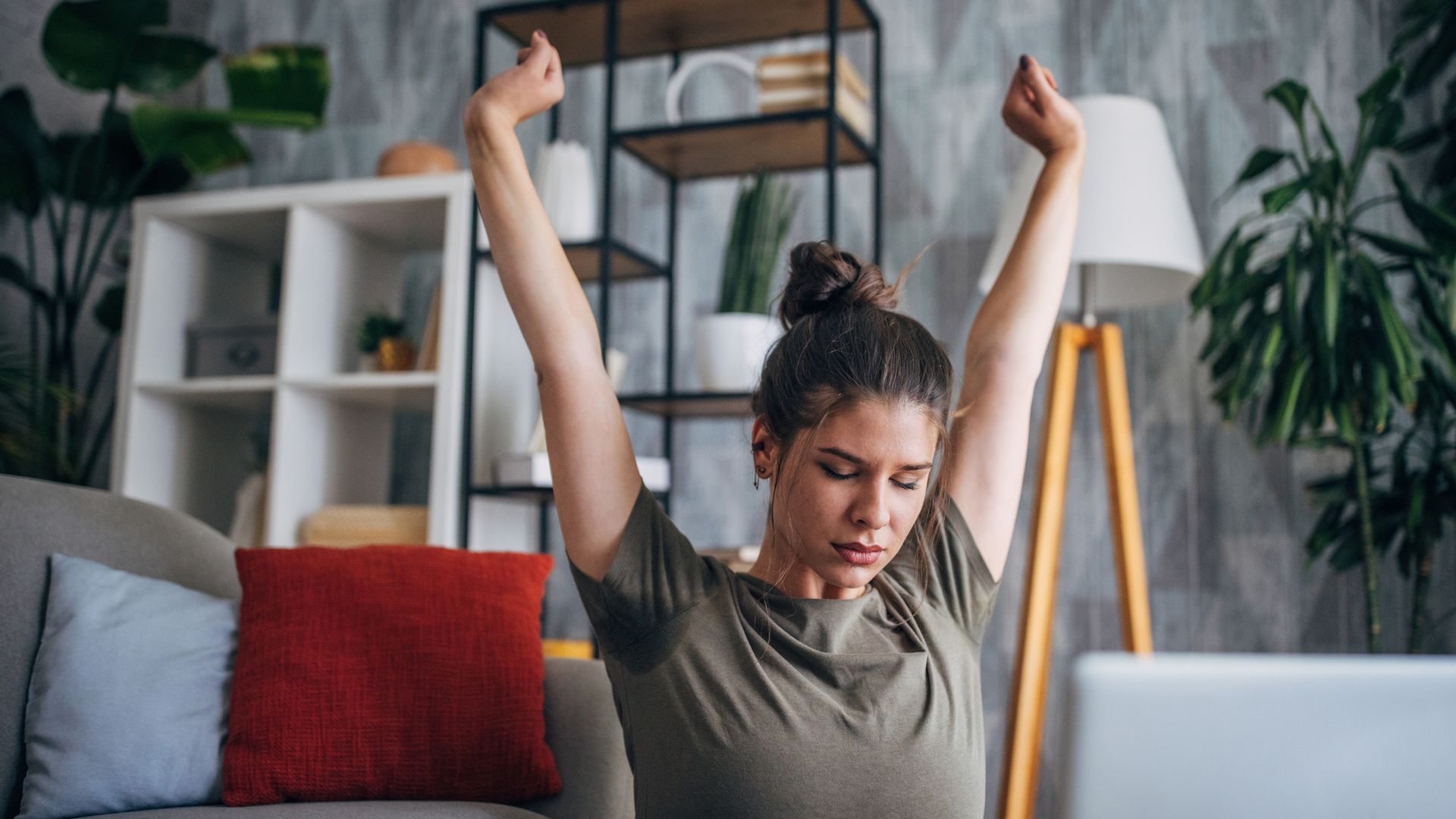 Mujer estirando los brazos hacia arriba al estar cansada, mientras está sentada frente a su ordenador portátil con una taza de café en la mesa; está sentada en el suelo del salón de su casa.