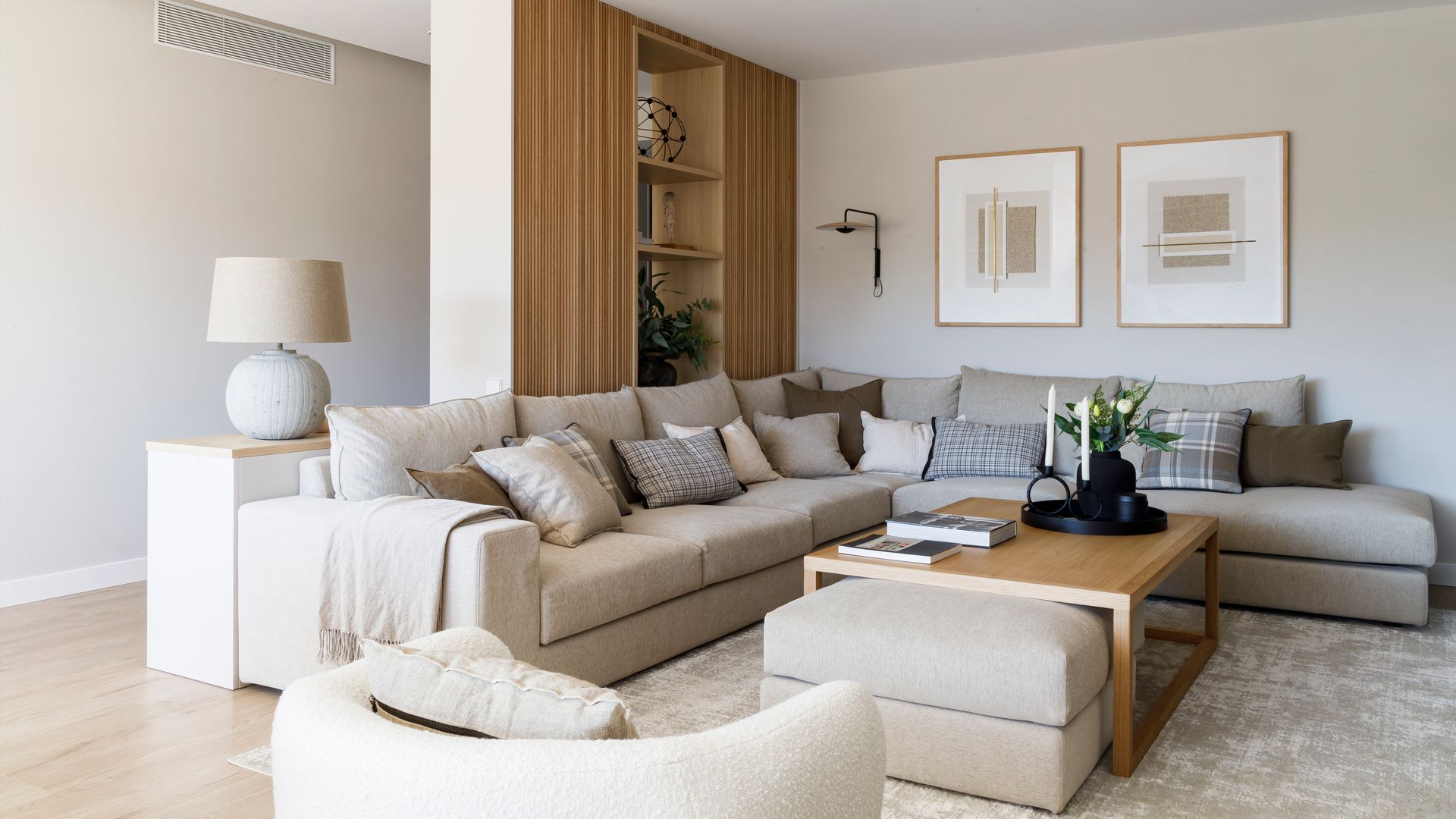 Salón con sofás beiges con chaise longue en uno de sus extremos, alfombra, estantería sin trasera que separa el pasillo del estar, cuadros en la pared, lámpara de sobremesa 