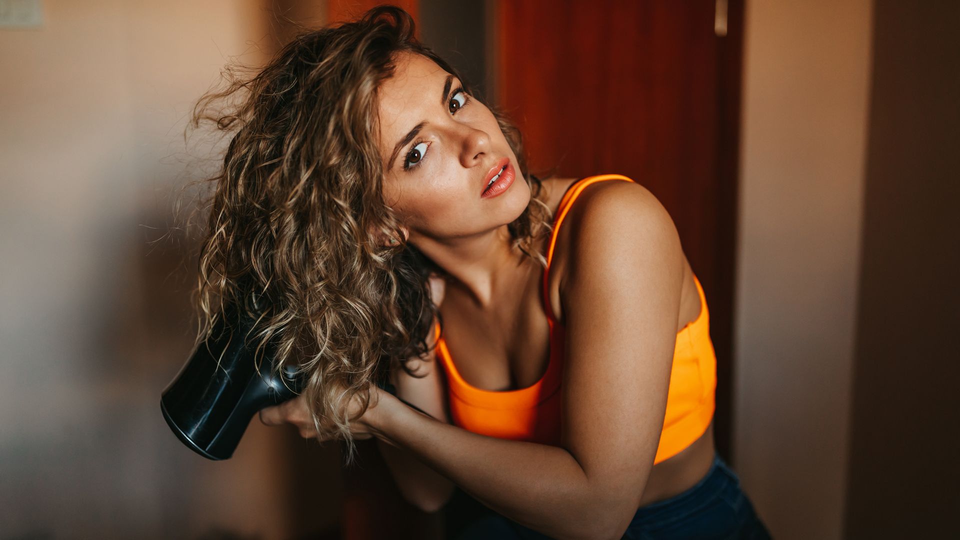 Mujer joven secándose el pelo con un difusor, mirando hacia delante. Tiene la piel bronceada y ropa de verano.
