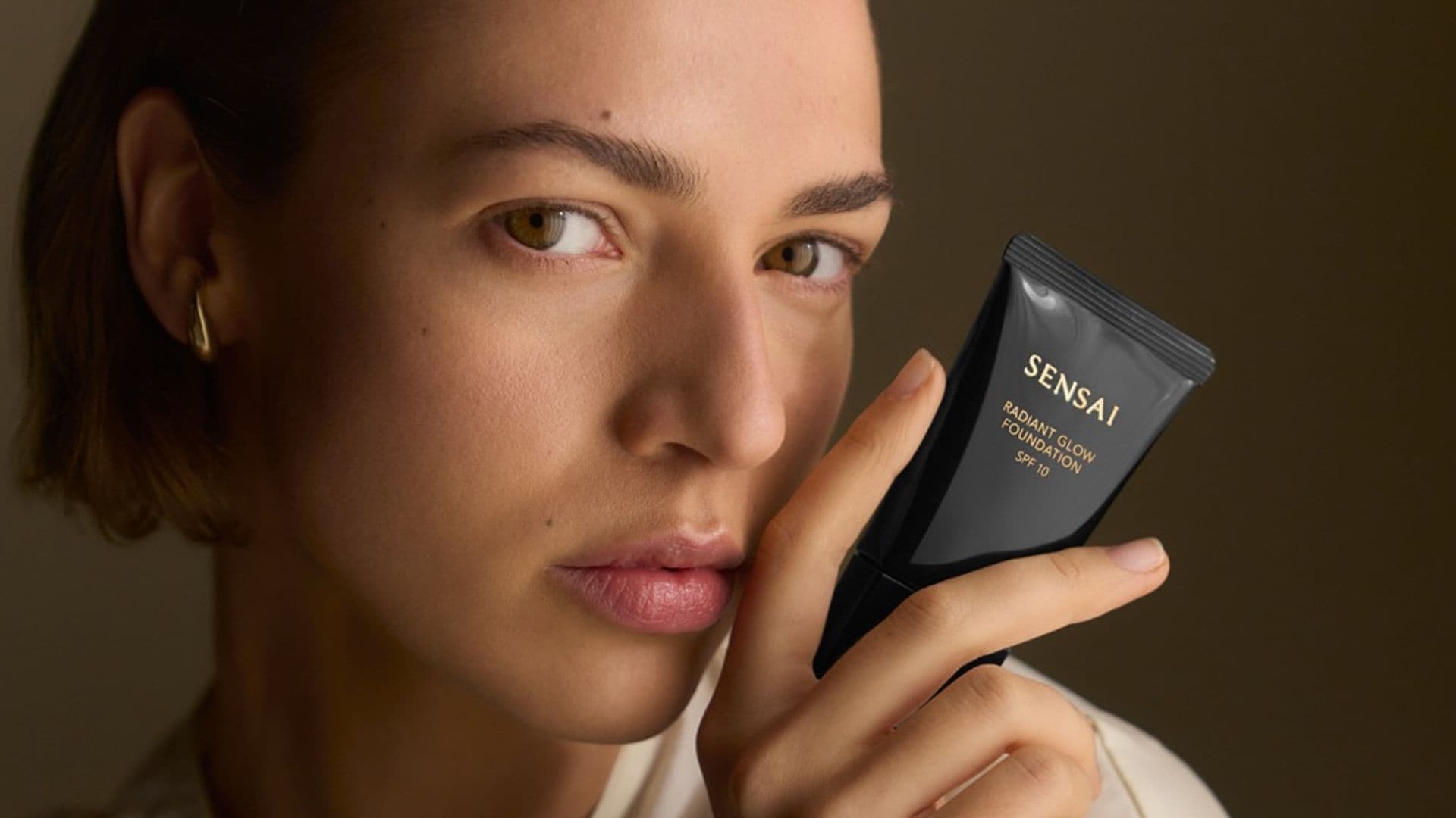 Rostro de mujer joven con pelo corto y blusa blanda mirando a cámara con base de maquillaje de Sensai en la mano