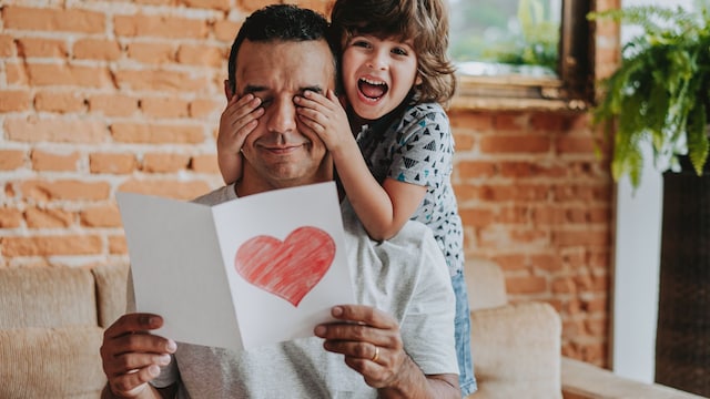 Padre sonriendo mientras su hijo le tapa los ojos y le entrega una tarjeta de felicitación con un corazón por el Día del Padre