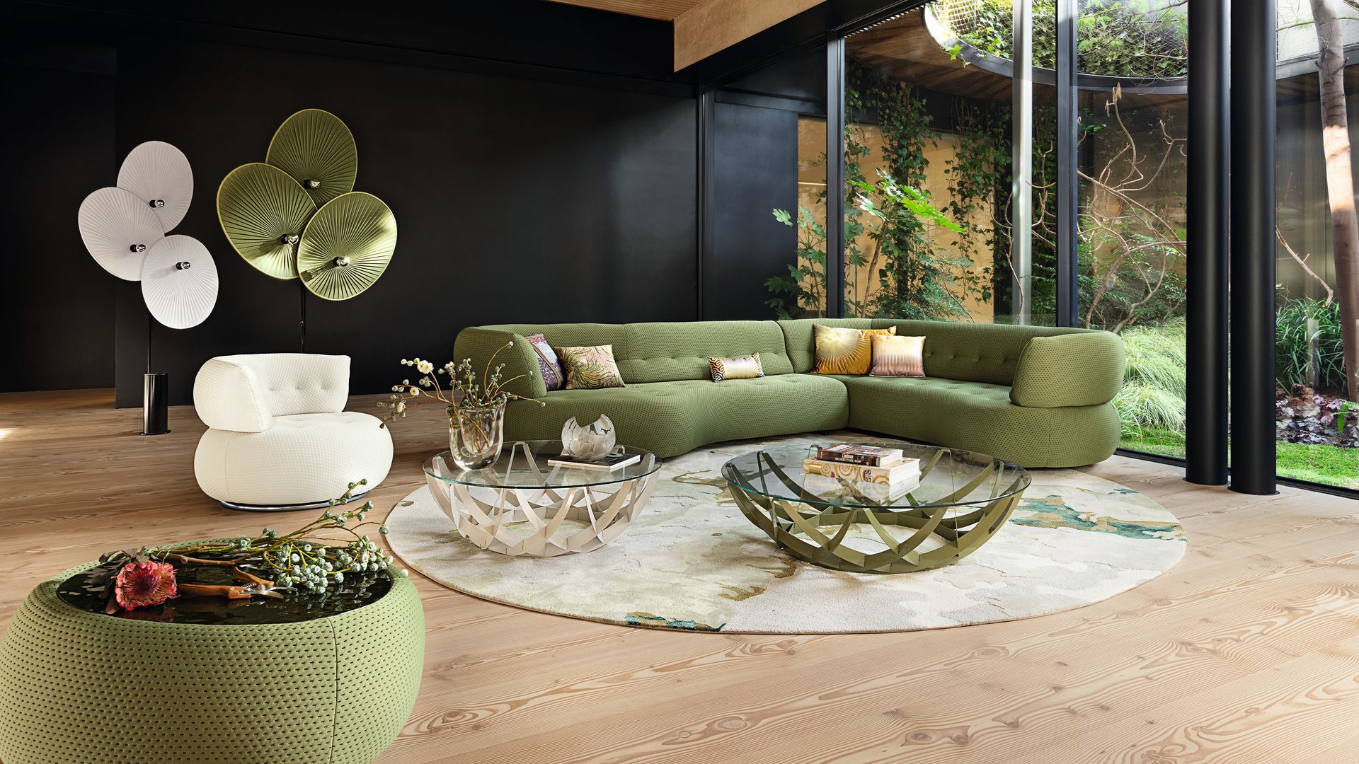 Salón con sofá verde con respaldo alto y en forma curva, con algo,bra redonda, grandes ventanales, dos mesas de centro con el sobre de cristal, adornos en pared negra