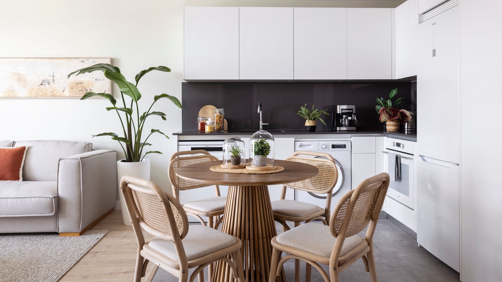 Cocina blanca, abierta con comedor, antepecho negro lámpara de techo, mesa de comedor redondo, sillas de rejilla, maceta XL