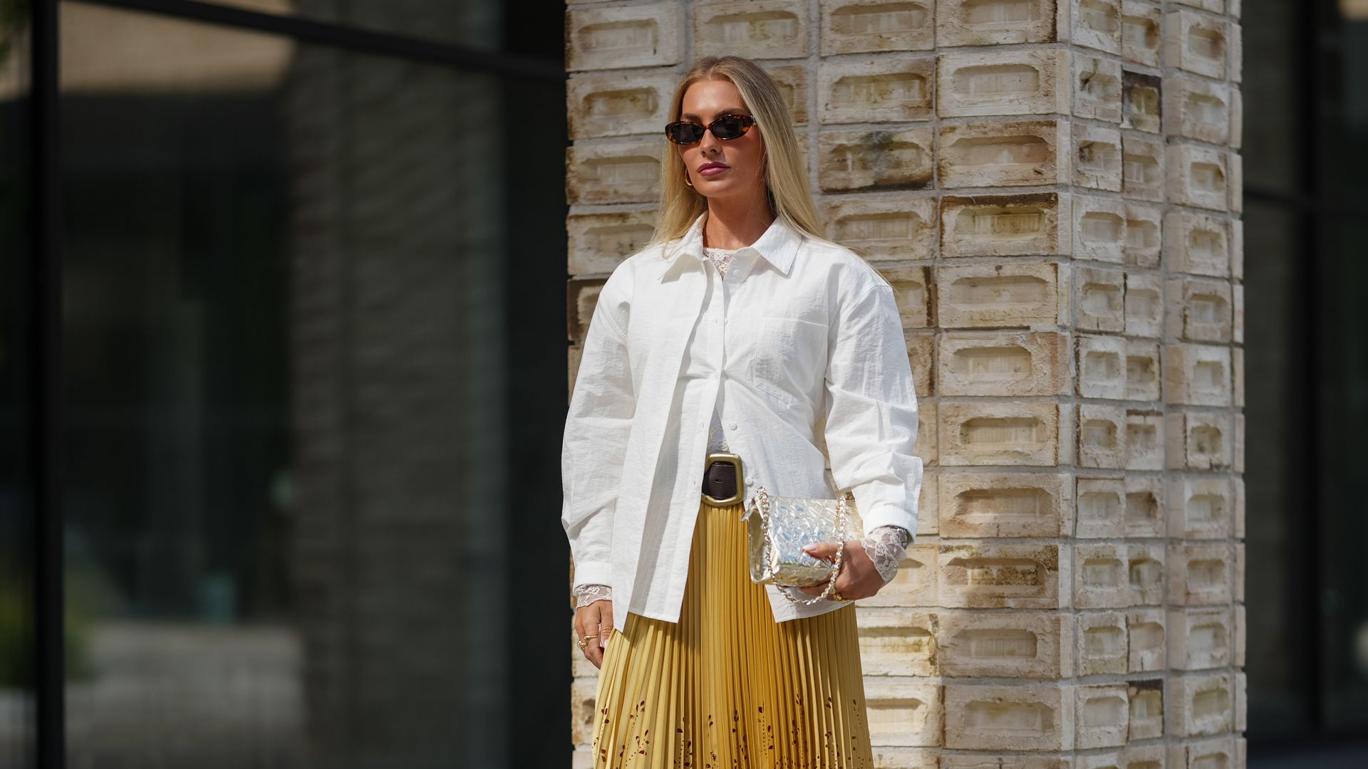 Street Style At Copenhagen Fashion Week SS26. Chica joven rubia posando con su camisa blanca, su falda plisada en amarillo y cinturón a la cintura, bolso de cadena en la mano izquierda y sandalias de tacón medio en plateado.