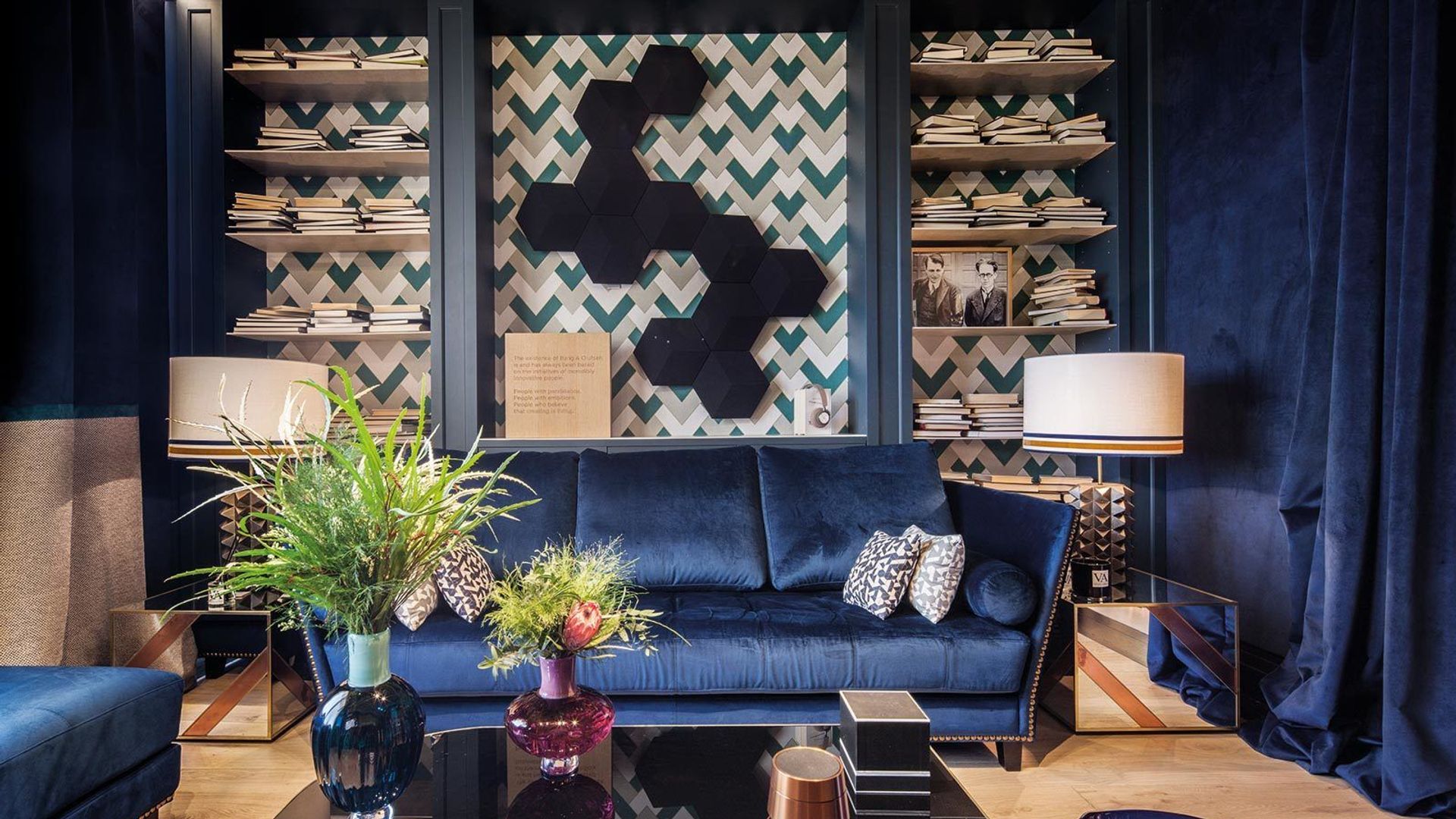Salón con sofá azul de terciopelo, cortinas a juego, mesa de centro con el sobre de cristal oscuro, librería a medida con papel pintado, libros, fotografías antiguas, lámparas e sobremesa, jarrones de cristal en azul y rosa