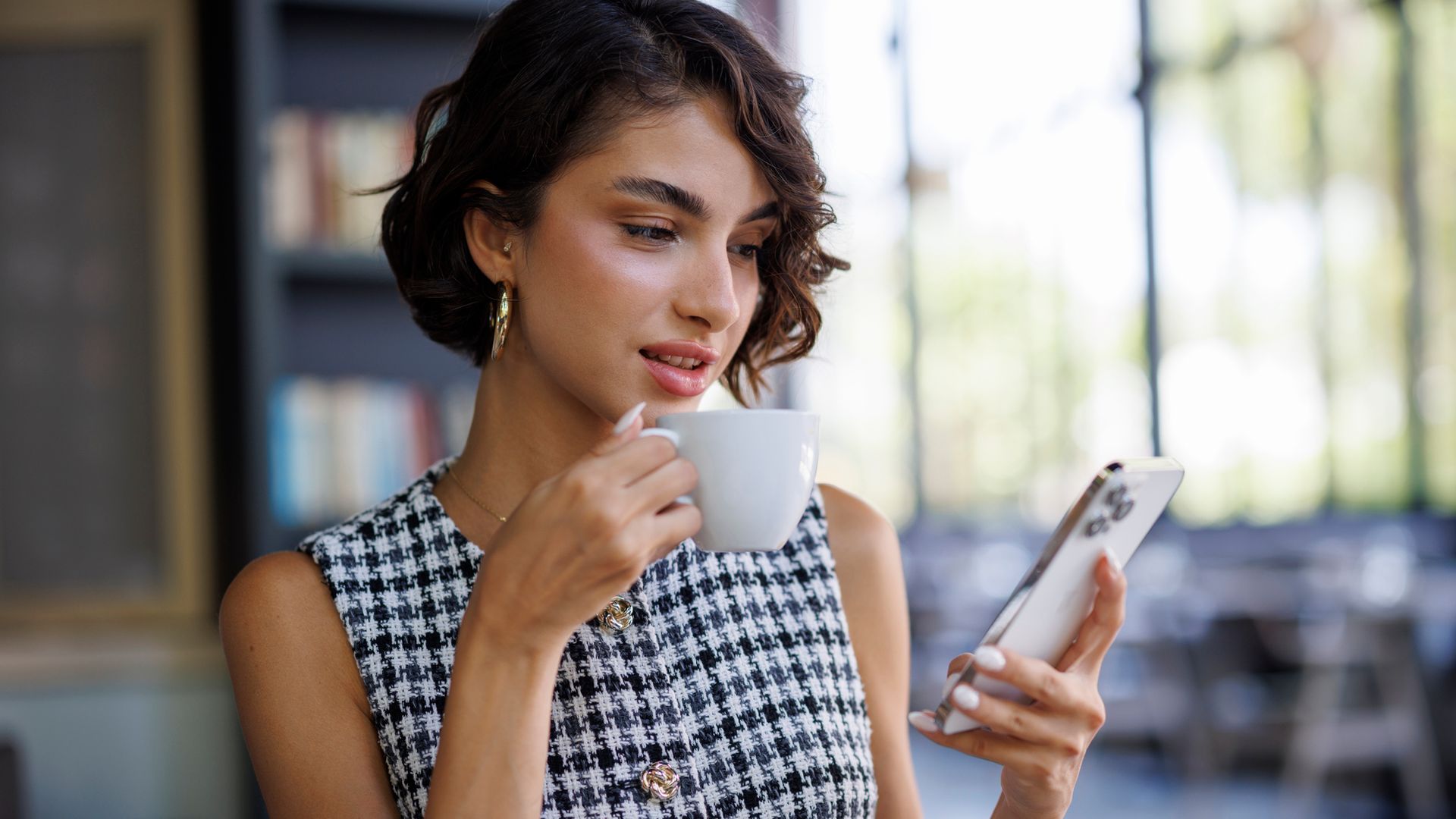Mujer joven y elegante con pelo corto rizado sentada en la terraza de una cafetería con chalecho vichy, tomándose un café y con el móvil en la mano