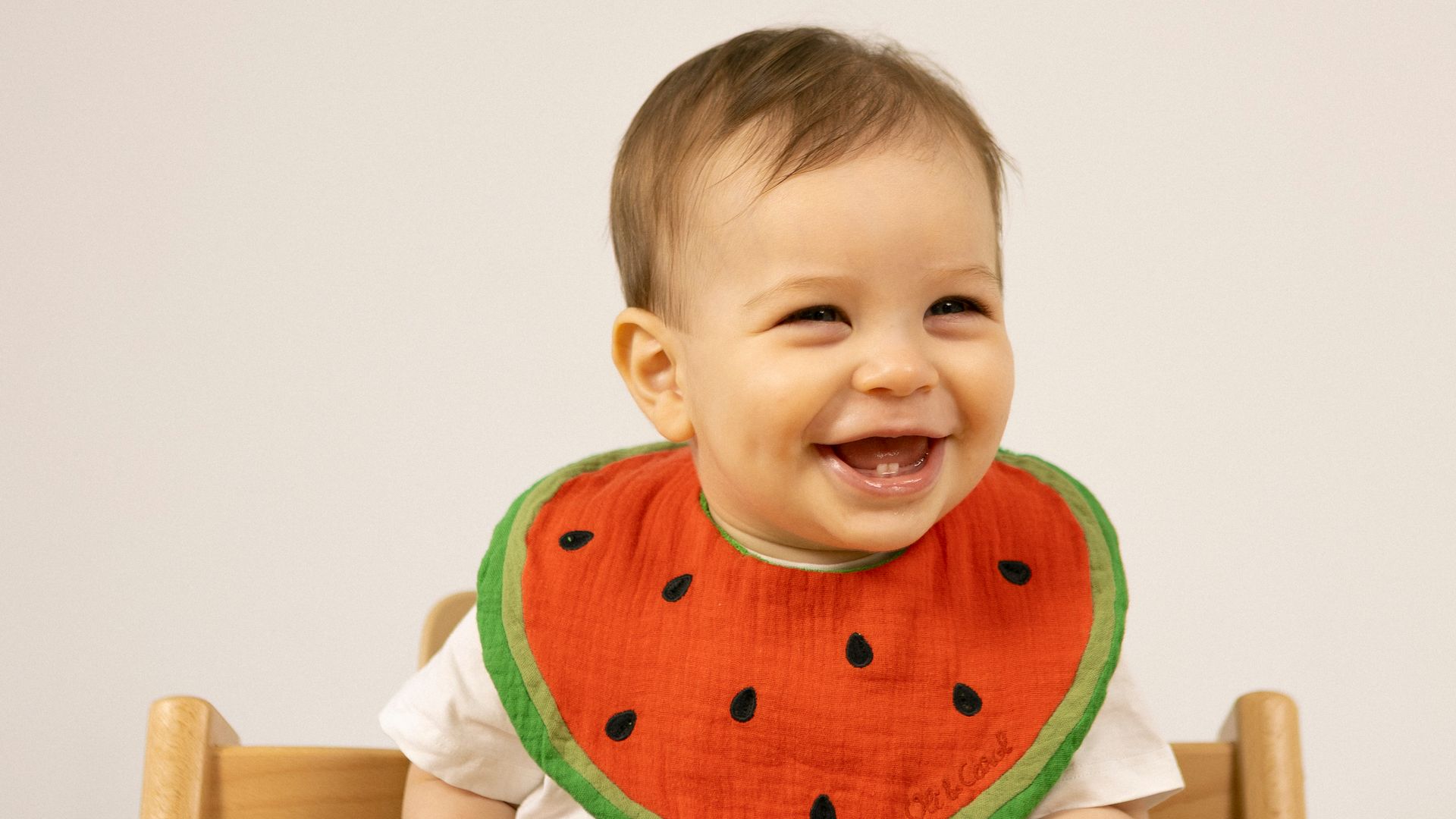 Bebé con un babero en forma de sandía y sentado en su trona espera, sonriente, su comida