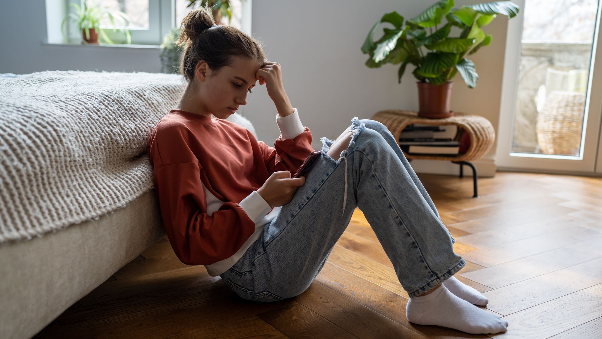adolescente preocupada mirando al móvil, sentada en el suelo de su habitación