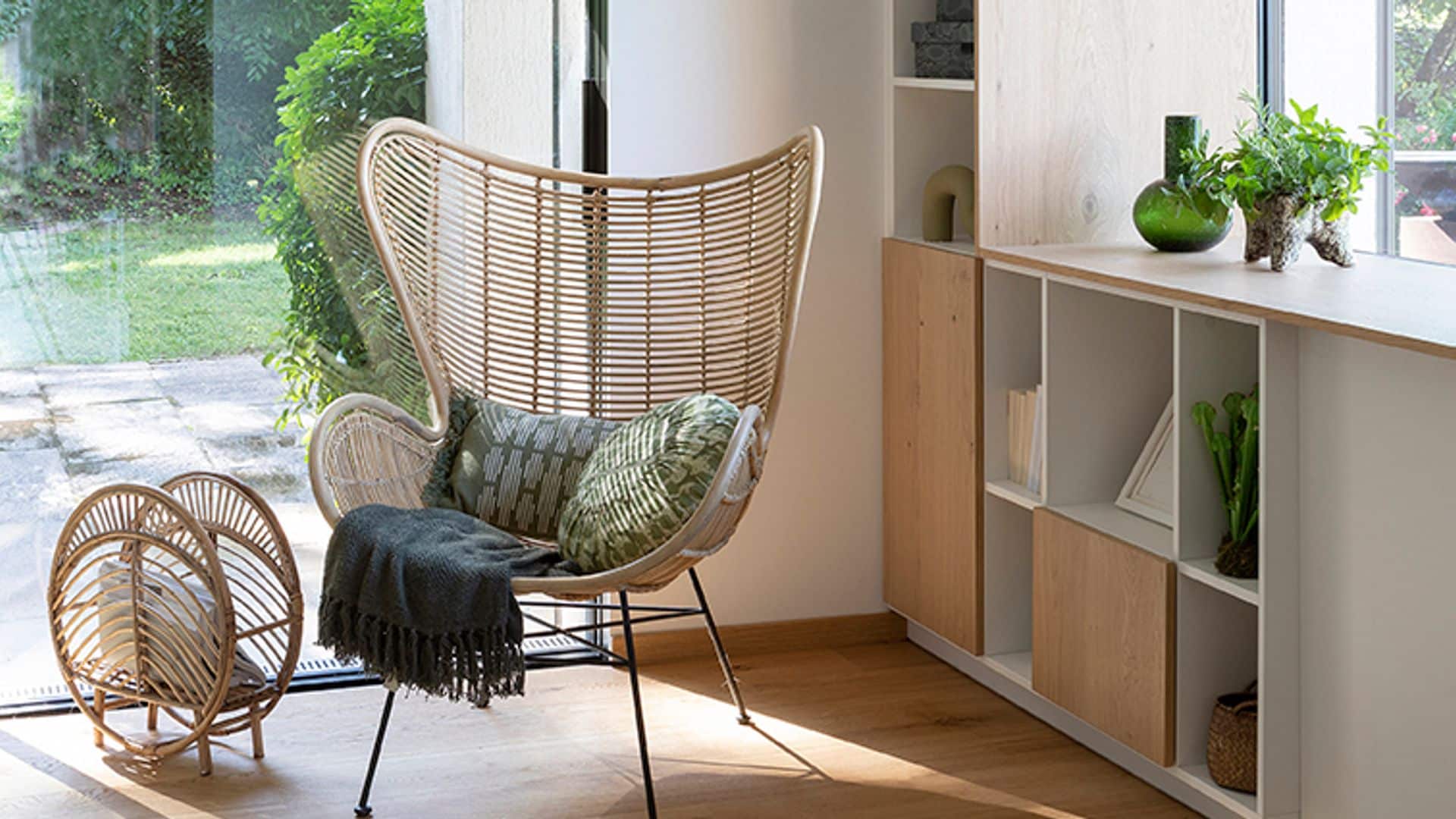 Rincón con estructura a medida rodeando la ventana con librería baja, plantas en el alféizar, suelo de madera, sillón de fibras vegetales, manta, cojines, revistero de fibras vegetales