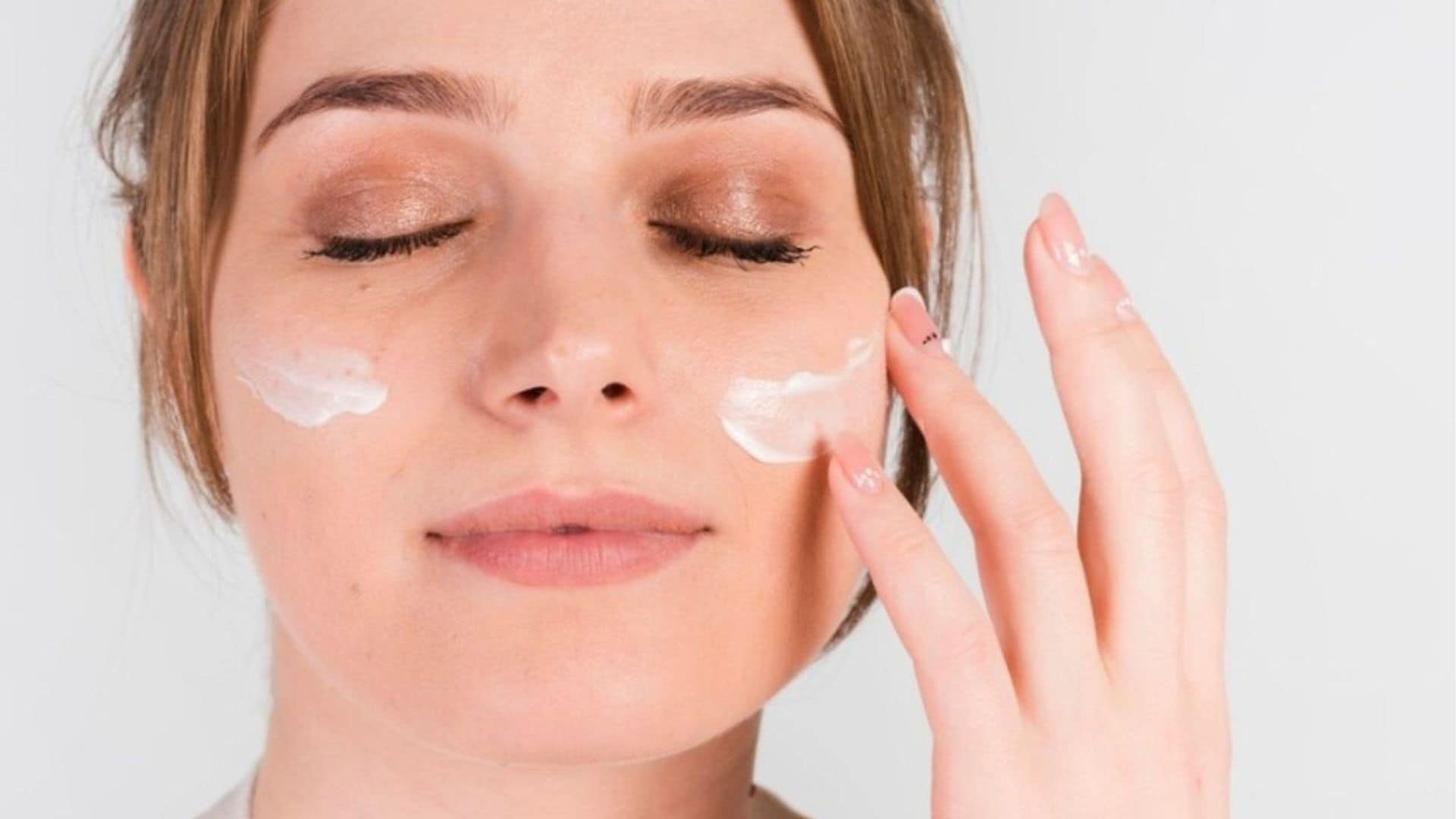 Chica joven echándose crema en la cara, con los ojos cerrados.