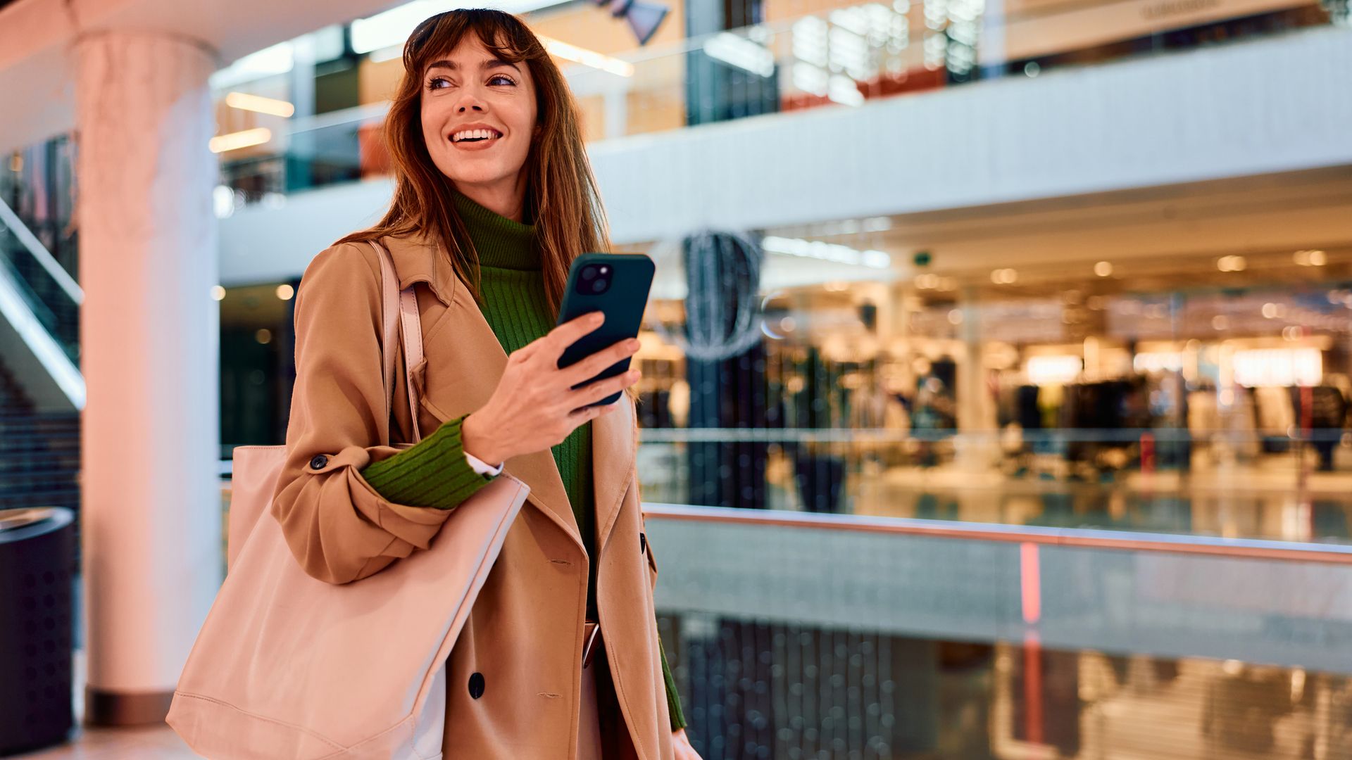 Mujer sonriendo en un centro comercial con el teléfono móvil en la mano y ropa de otoño-invierno.
