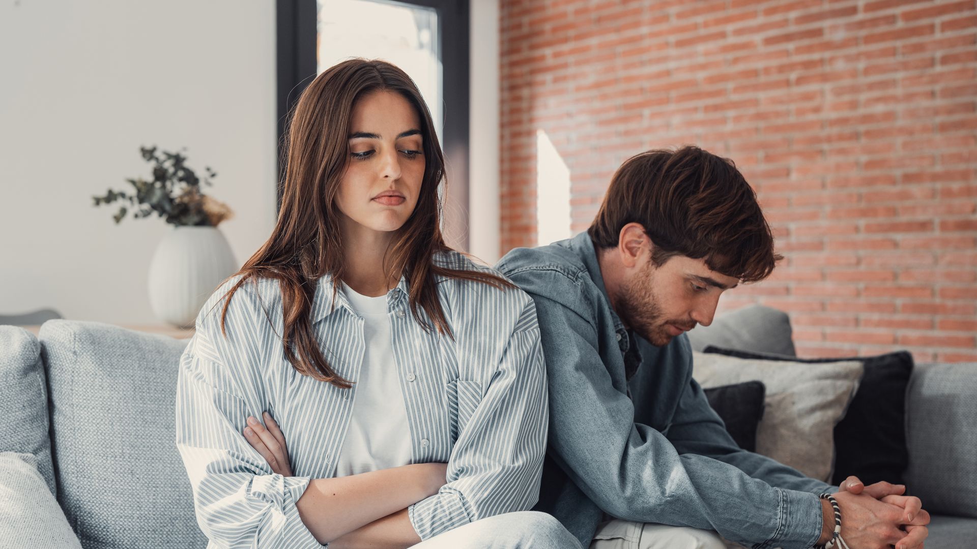 Mujer y hombre sentados en sofá, ella con brazos cruzados, él con manos entrelazadas.