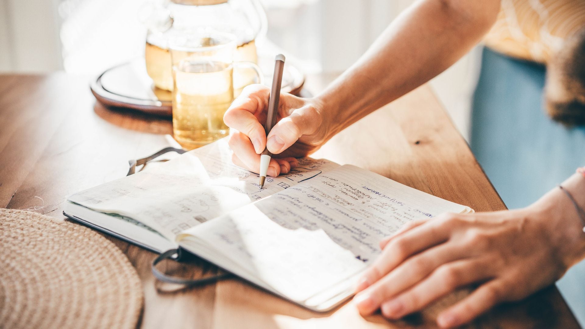 Mujer escribiendo en su agenda o cuaderno sobre una mesa, con una tetera e infusión detrás. Plano detalle del cuaderno y las manos.