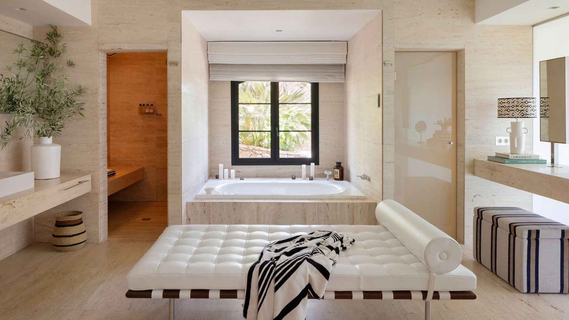 Baño con bañera de hidromasaje, ventana, estor, zona de tocador con puff tapizado, cama relax de cuero blanco
