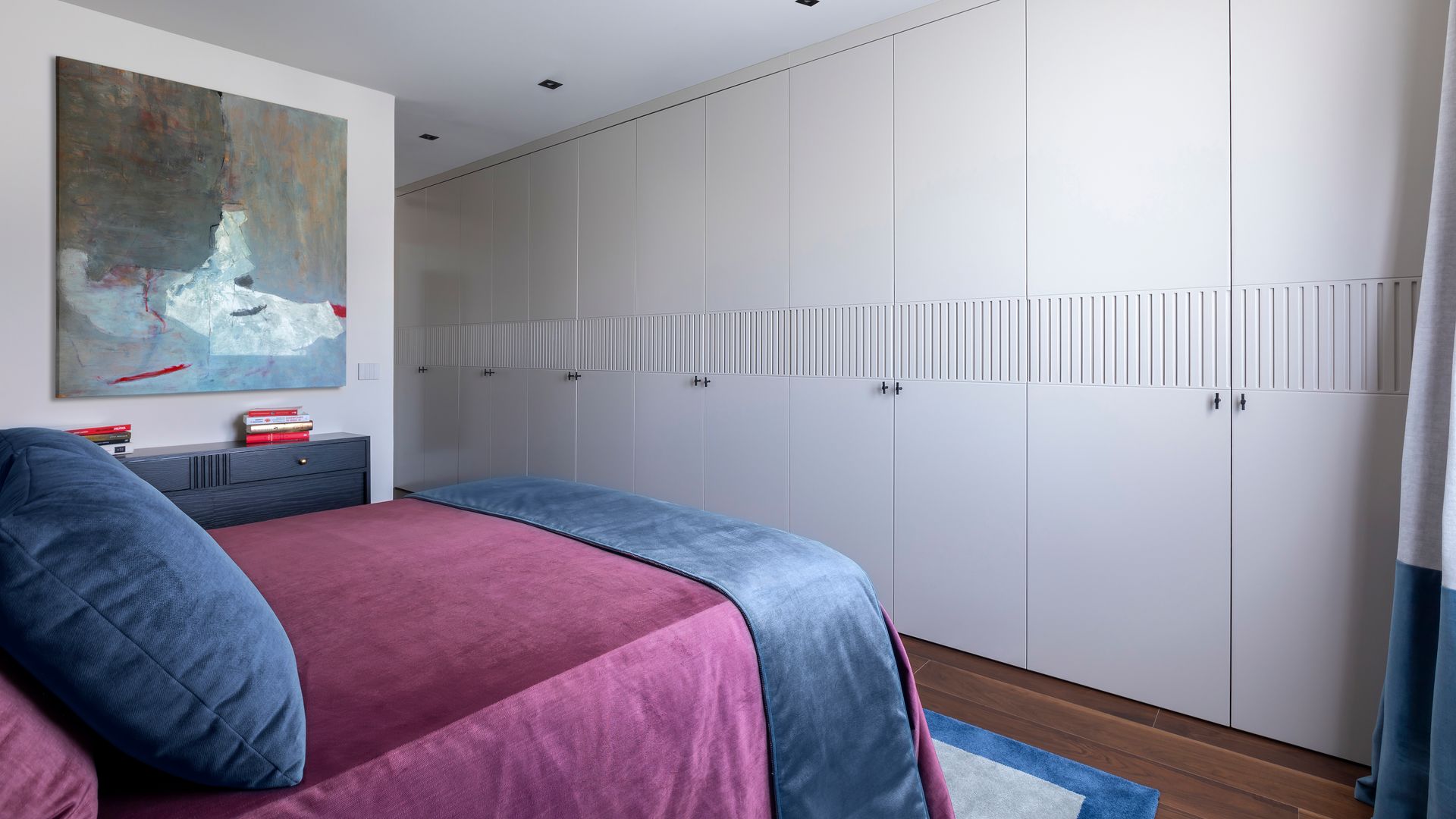 Dormitorio de matrimonio con pared de armarios blancos frente a la cama vestida con risa de cama burdeos y azul, alfombra, suelo de madera y cuadro 