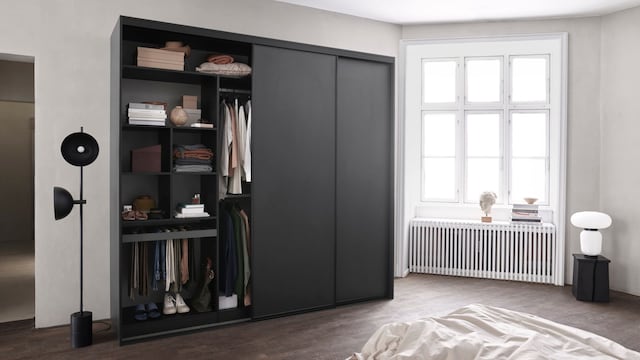 Dormitorio con gran ventanal, radiador antiguo blanco, suelo de madera oscuro, armario exento negro abierto con baldas y pantaloneros, lámpara de pie negra 