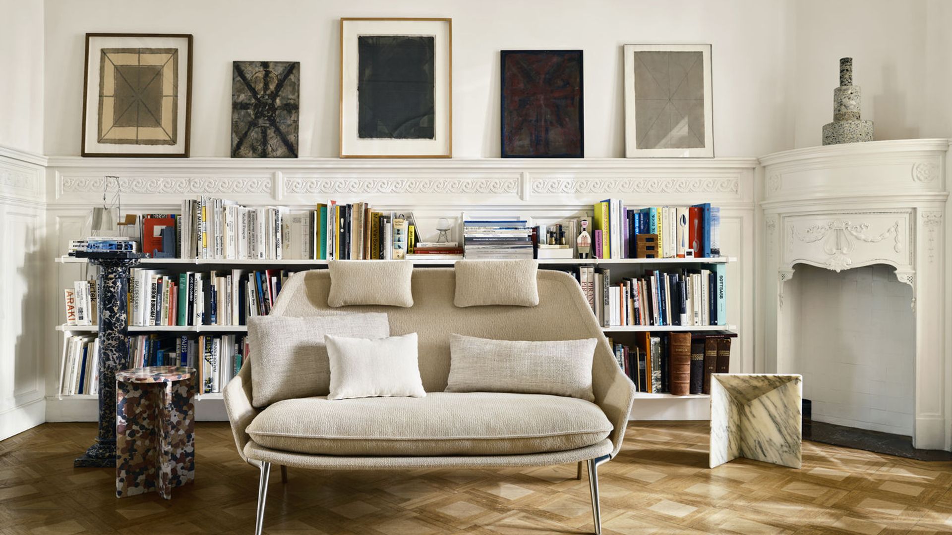 Salón con chimenea, sofá beige, pared detrás del sofá con estantería, balda y cuadros, suelo de madera 