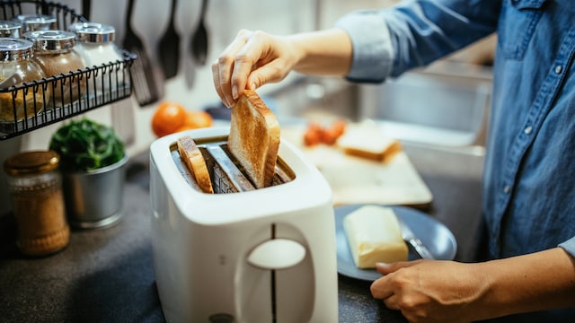 Mujer cogiendo una tostada recién salida del tostador en la cocina, con mantequilla y tomate en la encimera, preparada para desayunar