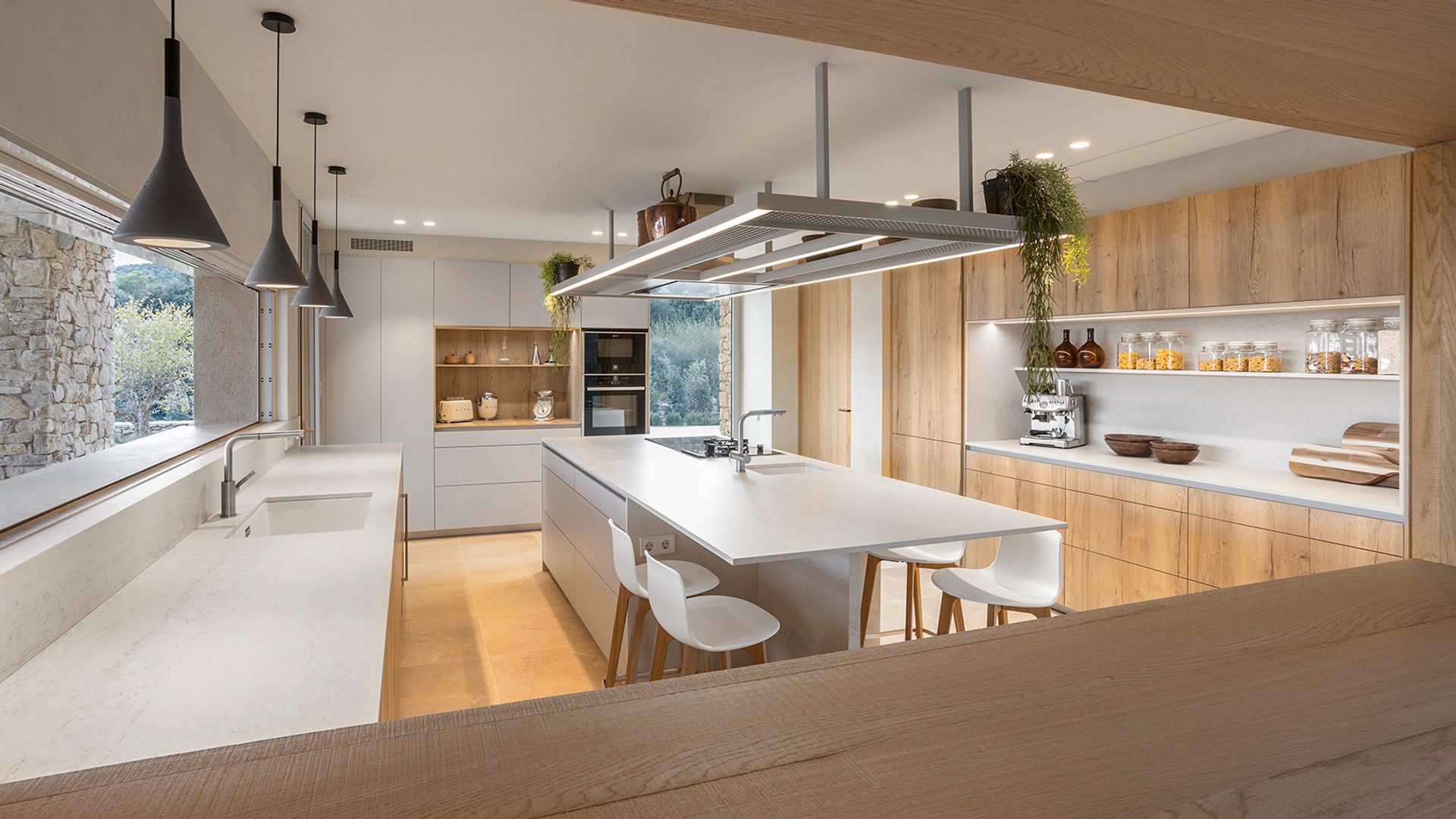 Cocina grande con gran ventanal, con muebles blancos y de madera, isla central con zona de fuegos y fregadero, sobre volado en la isla para desayunos con taburetes, campana de techo 