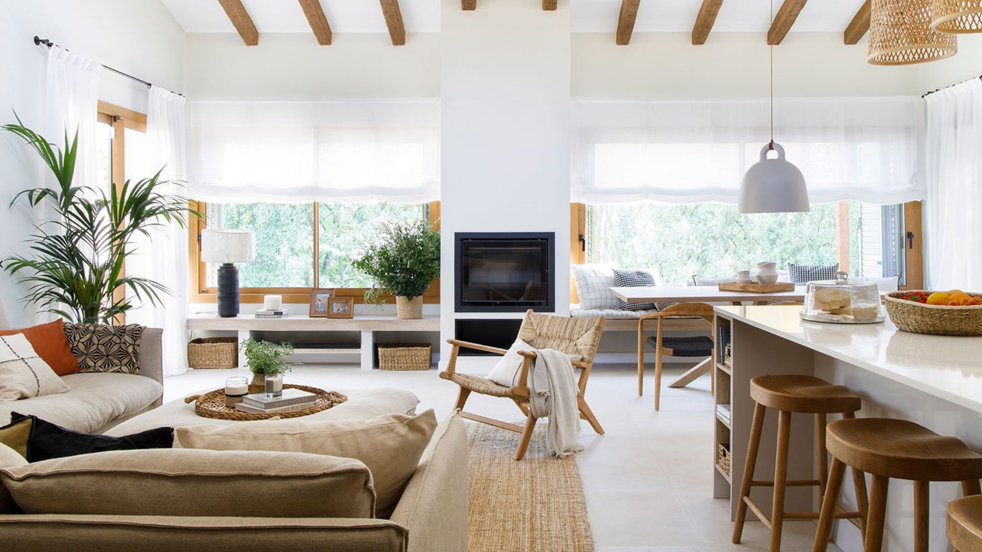 Salón comedor y cocina con isla y taburetes, vigas de madera, banco bajo la ventana, alfombra de fibras vegetales, mesa de centro tapizada