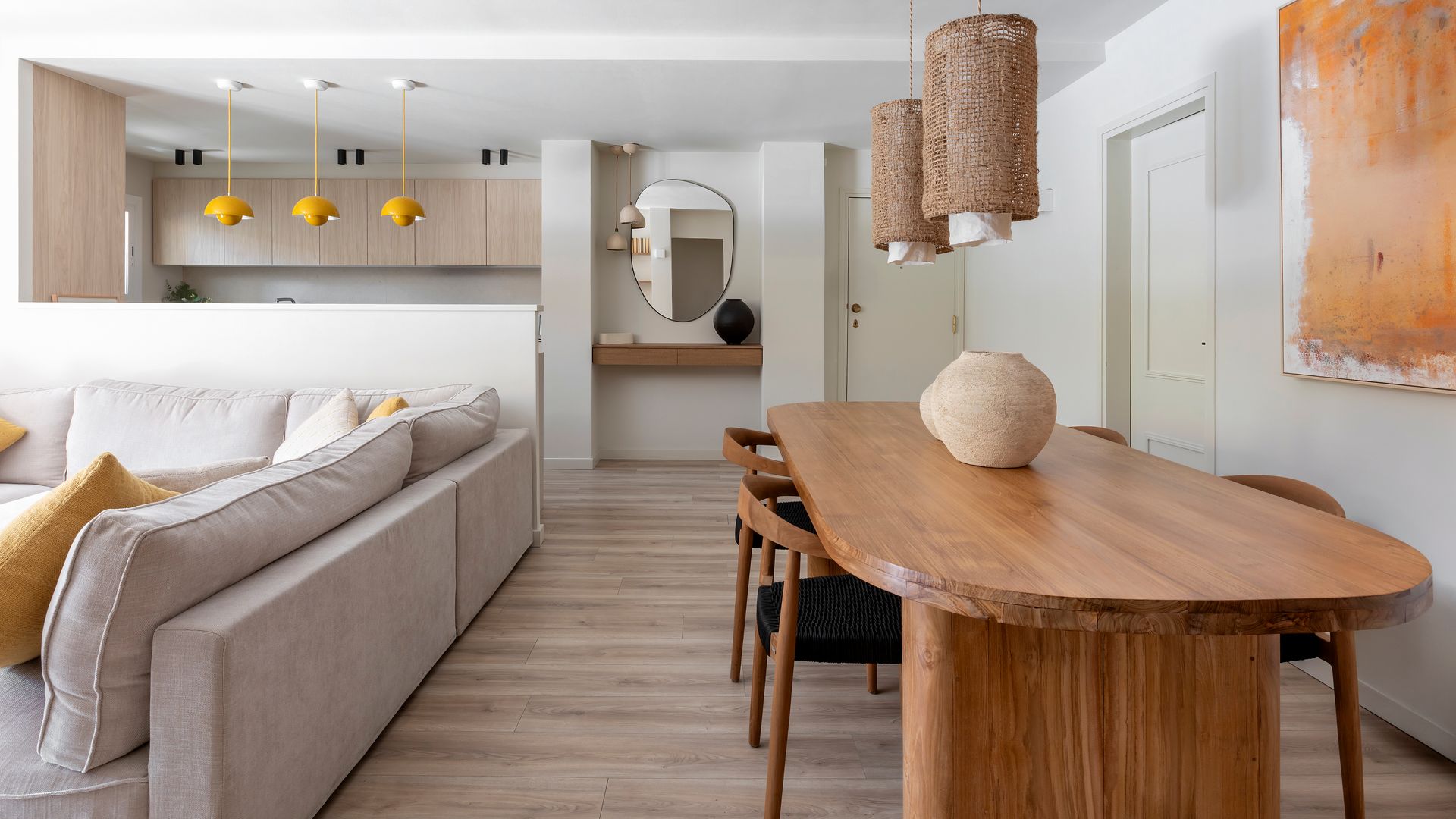 Salón comedor y cocina con mesa de comedor de madera, ovalada y XL, sillas de madera con el asiento tapizado, sofá gris claro, medio murete de obra separando la cocina del salón comedor