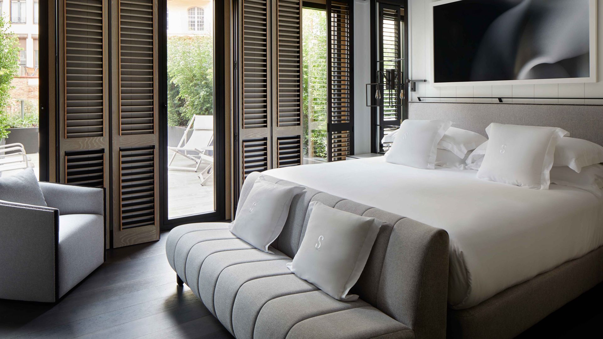 Dormitorio con salida a la terraza con contraventanas de madera, cama con sábanas blancas, cuadro en la pared del cabecero, cabecero tapizado gris, banco a los pies de la cama 