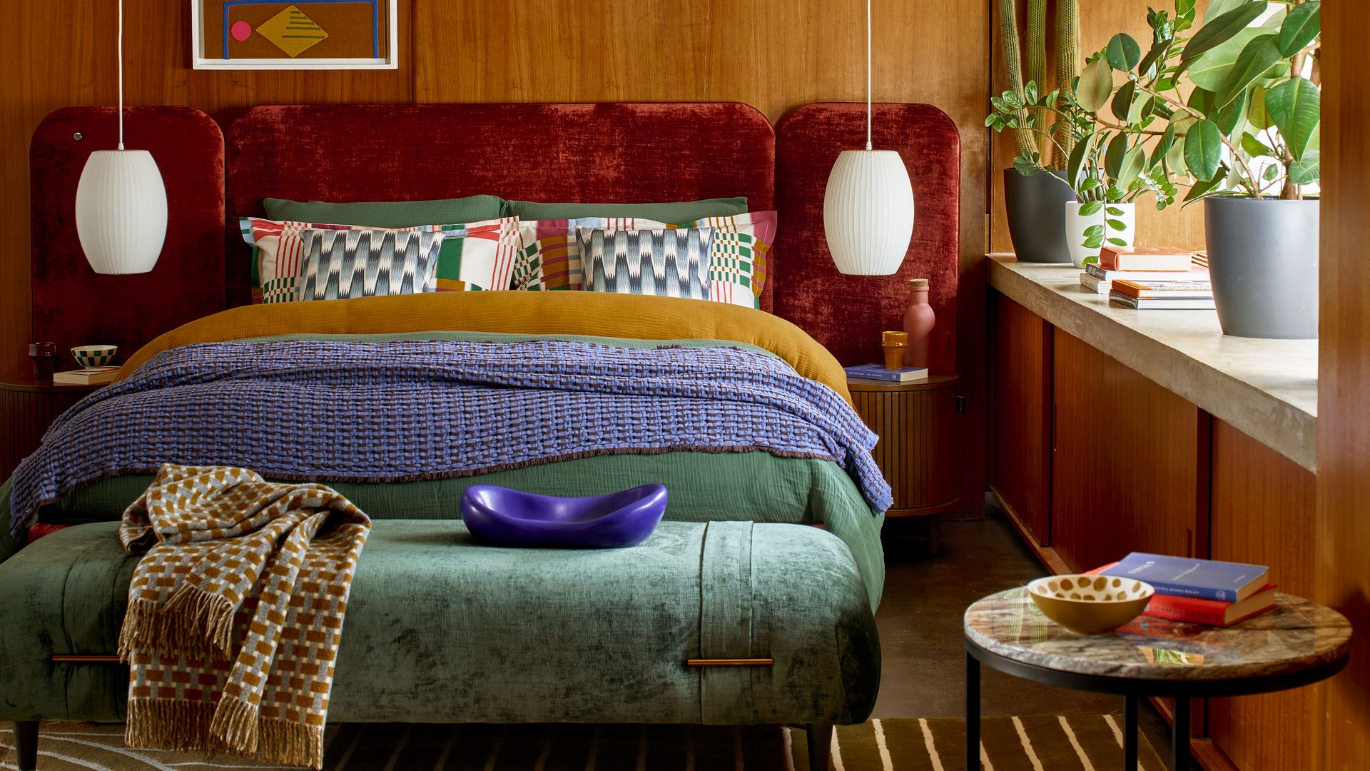 Dormitorio con pared en madera, cabecero tapizado en color marrón oscuro, banqueta tapizada en verde, plantas en la ventana, mantas sobre la cama