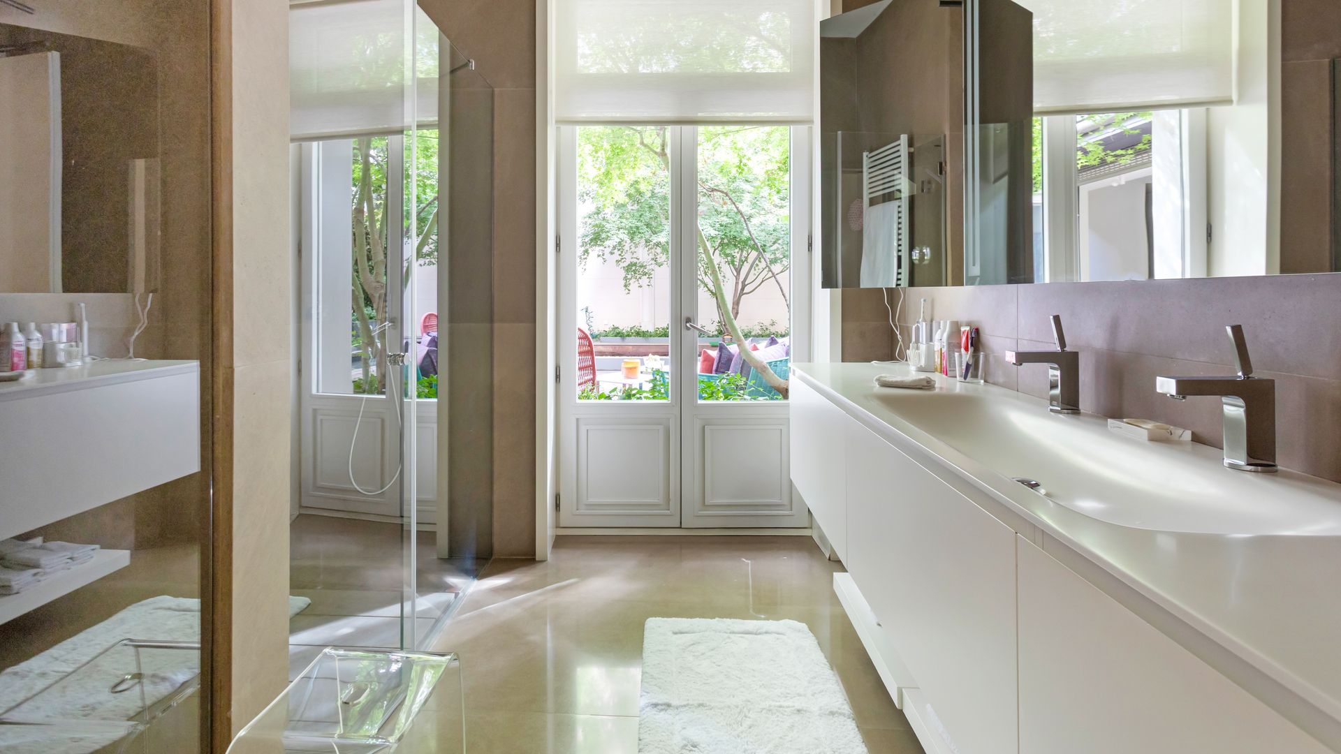 Baño con salida a la terraza, ventanas de madera, encimera blanca, lavado con dos grifos, estores