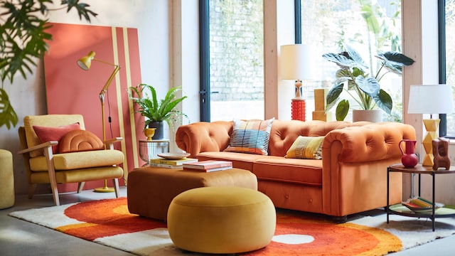 Salón con sofá cama cerrado en color naranja, mesa de centro tapizada, alfombra naranja y blanca, butaca y puff