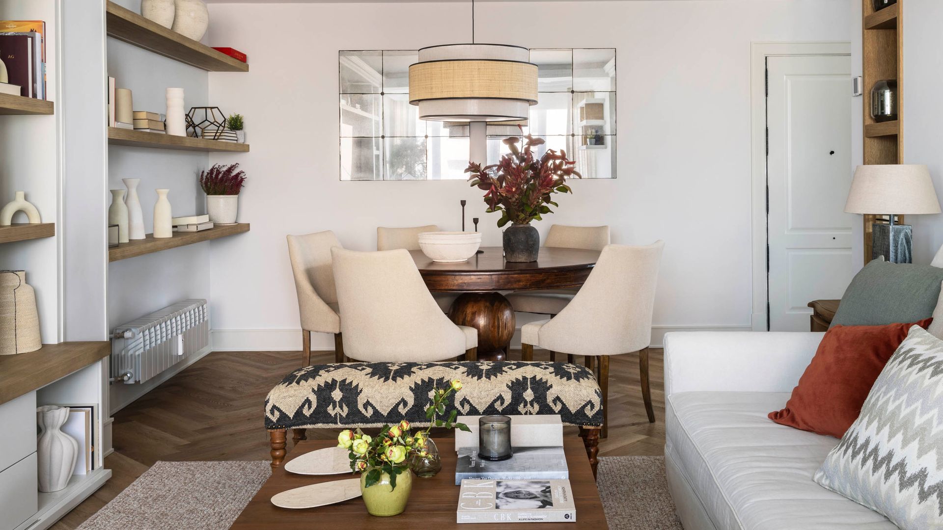 Salón comedor con sofá blanco, molduras en el techo, espejo al lado de la mesa de comedor, mesa de comedor redonda, mesa de centro de madera, sofá con cojines, librería a medida 