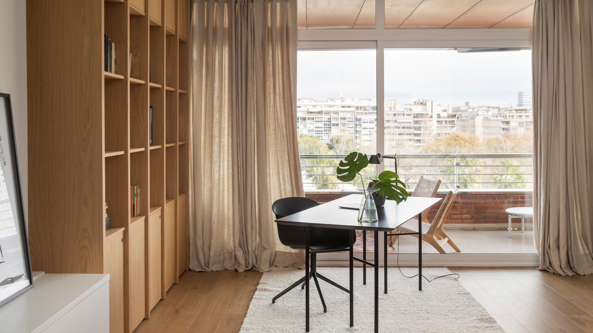 Despacho con estantería de madera, salida a la terraza, mesa de despacho negra, silla negra, alfombra blanca, cortinas beiges