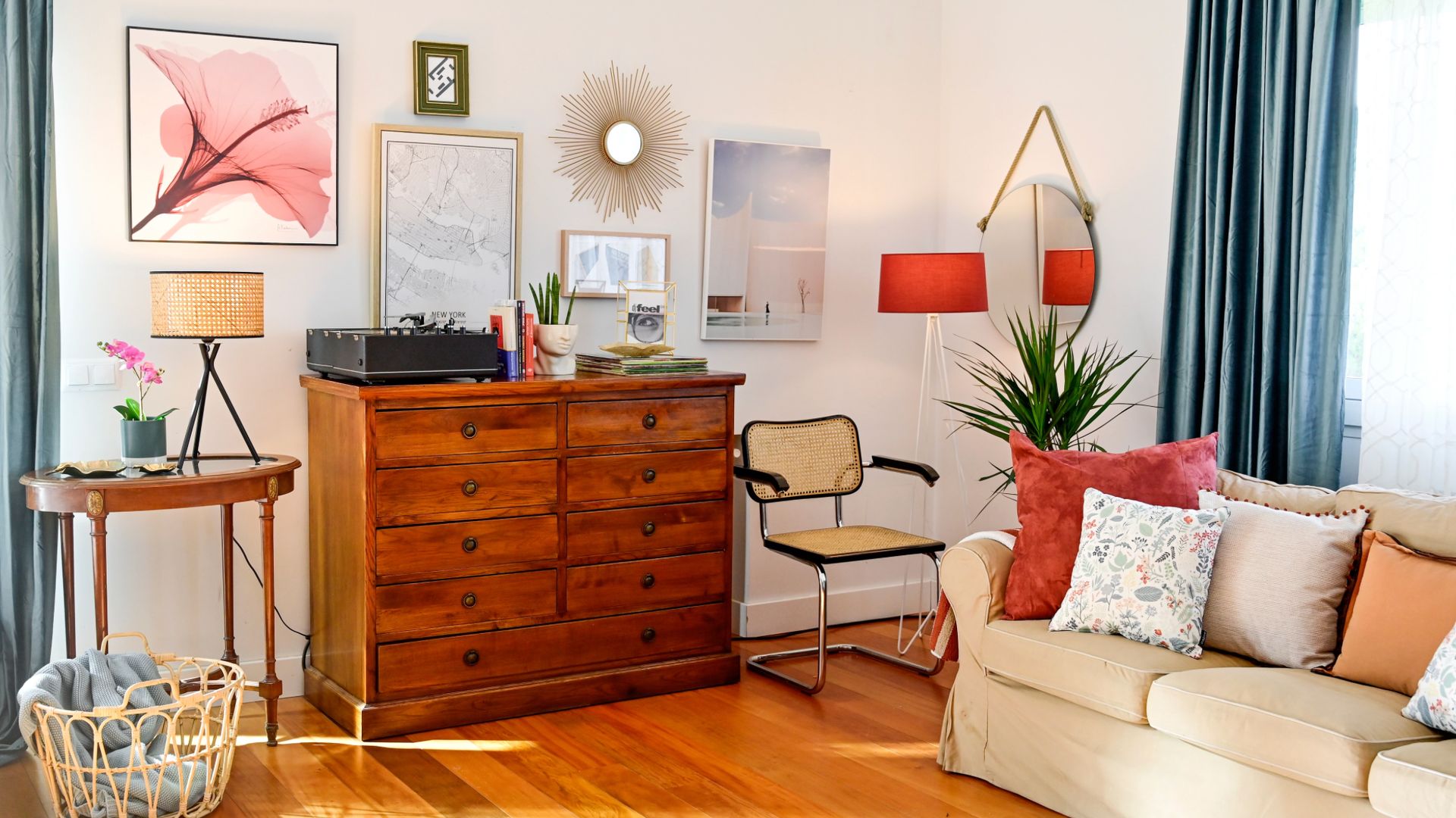 Salón con aparador de madera, tocadiscos, mesa auxiliar de madera, silla de rejilla, cuadros, lámpara de pie roja, lámpara de sobremesa, cojines, sofá beige