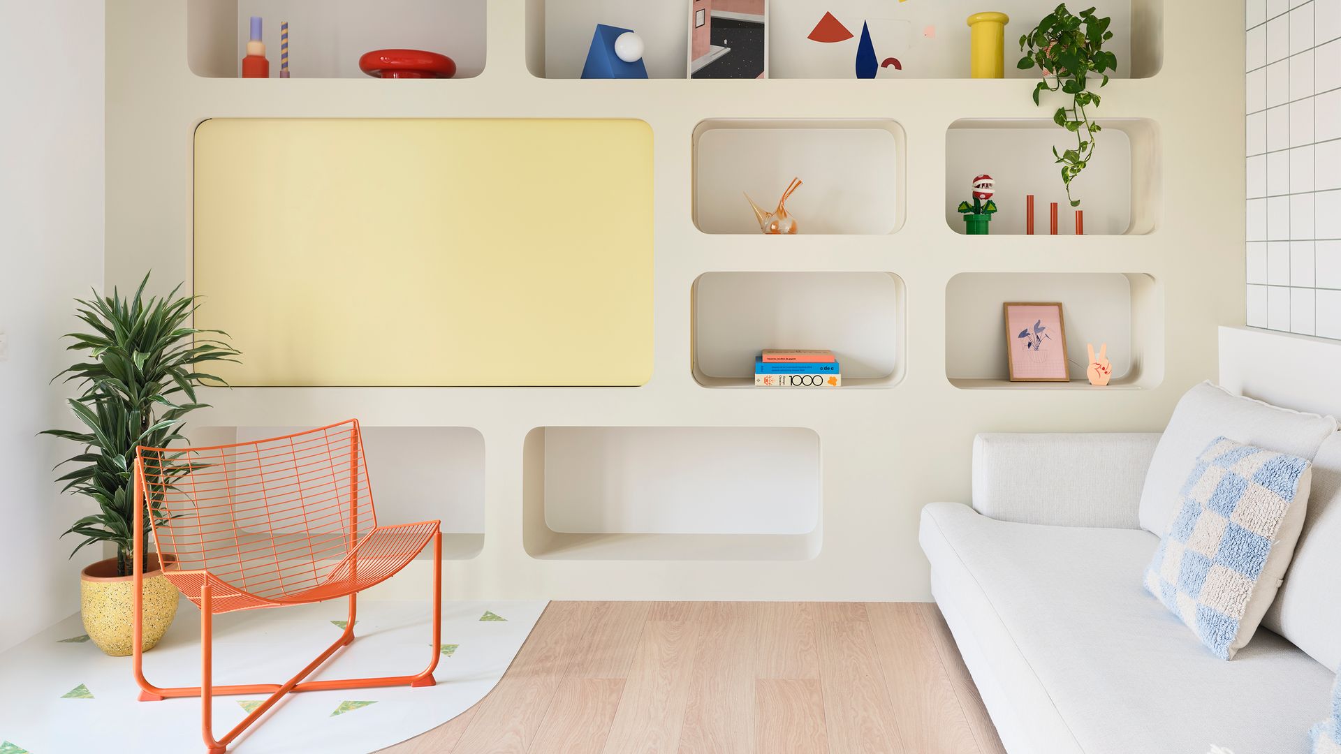 Salón con librería a medida, estructura amarilla que oculta la televisión, suelo de madera, sofá blanco, butaca metálica naranja, cojín azul cielo, adornos de colores en la estantería, plantas colgantes 