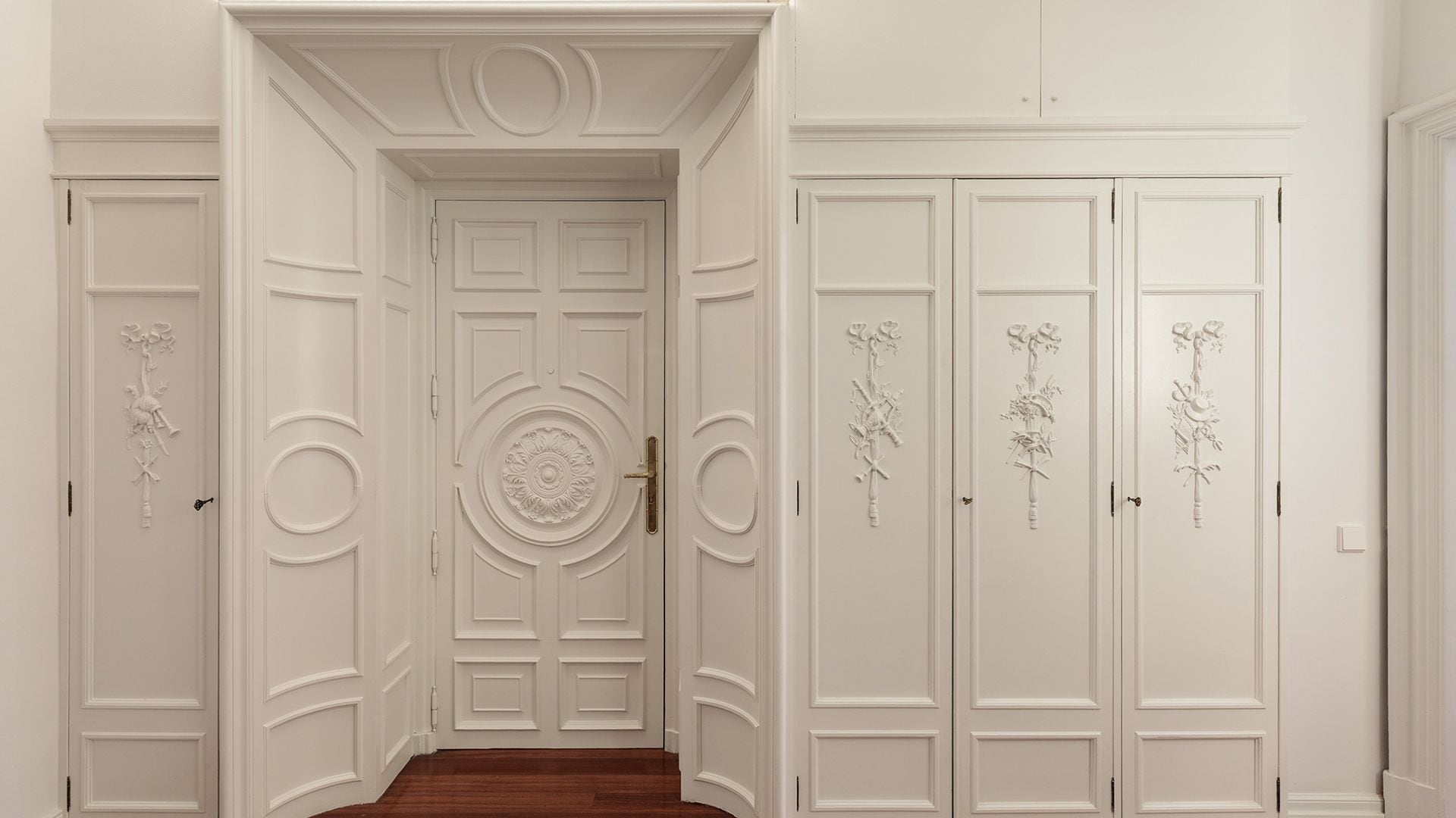 Entrada casa con paredes y armarios con molduras blancas y rosetones, suelo de madera oscura 