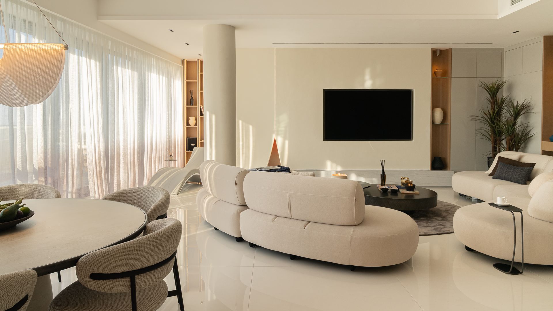 Salón comedor con sofás curvos blancos, columna, televisión colgada en la pared, mueble de comedor ovalada blanca, sillas tapizadas, suelo blanco roto