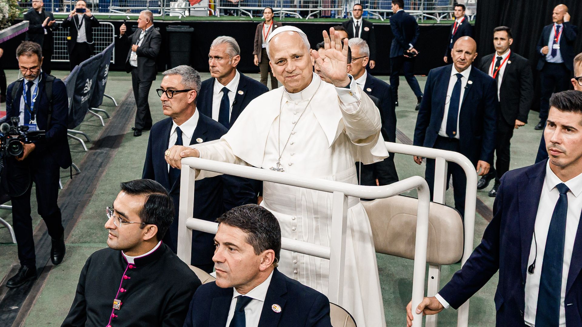 Papa Francisco saluda a la multitud en un estadio, acompañado por varios sacerdotes y oficiales.