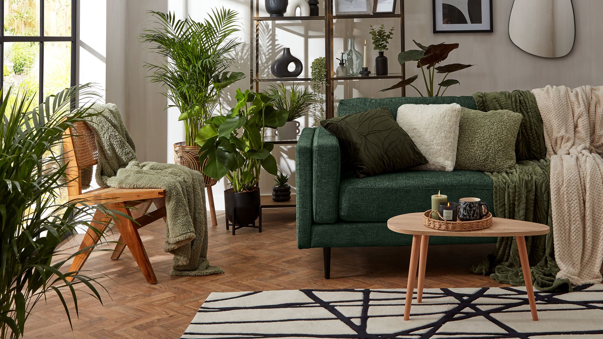 Salón con gran ventanal, sofá de color verde oscuro, plantas en el suelo, alfombra, cojines, mesa de centro de madera con bandeja de fibras vegetales con velas, mantas en el sofá, silla de rejilla, estantería con jarrones, marcos y adornos
