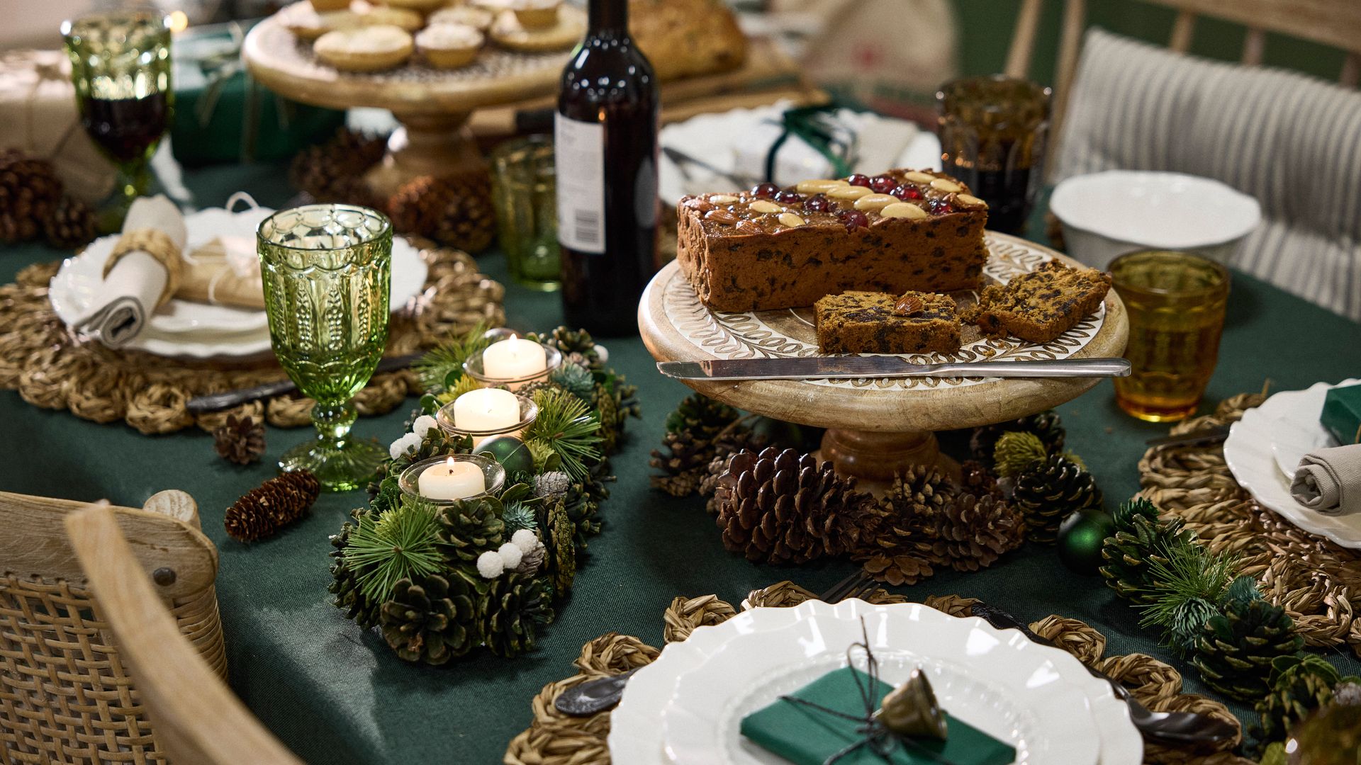 Mesa de Navidad con vajilla blanca, mantel verde, centro de mesa de piñas y velas, piñas sobre el mantel, botella de vino