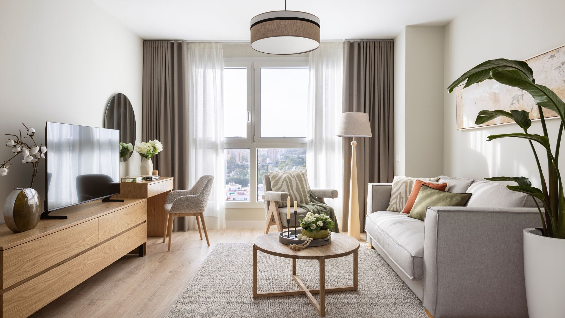 Salón con sofá cama, mueble de televisión de madera, mesa de comedor redonda, zona de escritorio, lámpara de techo, alfombra gris, cortinas y visillos, maceta grande en el suelo 