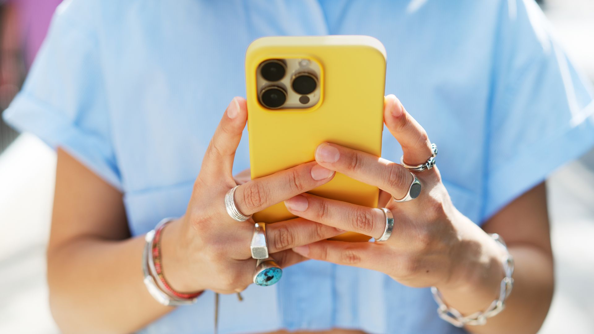 Mujer joven con teléfono móvil entre las manos, en un plano detalle de ellas con la cámara cerca por delante. El teléfono móvil es amarillo y las manos de la chica están llenas de anillos con ropa primavera-verano.