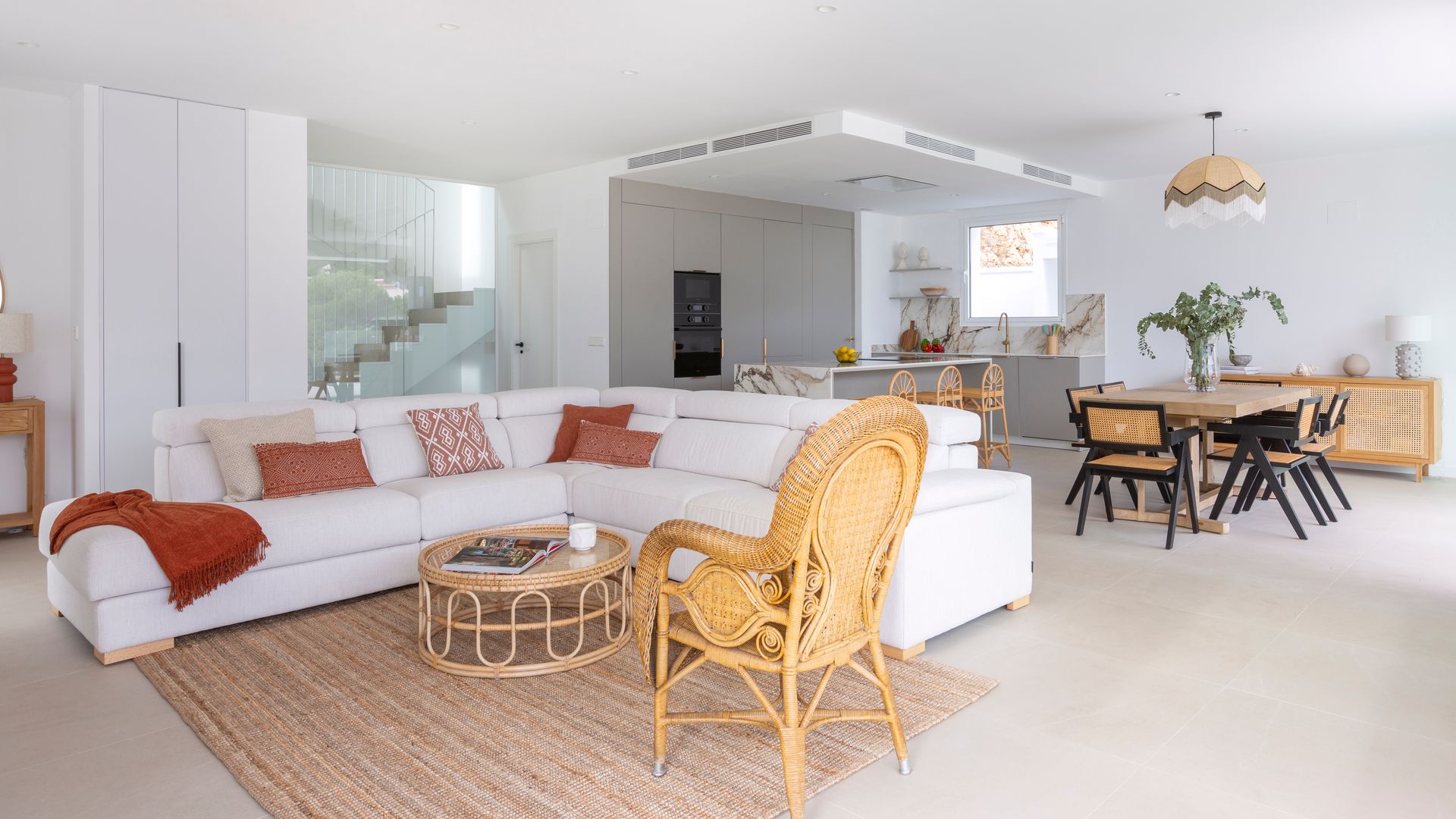 Salón comedor y cocina con sofá blanco en el centro, alfombra, mesa y butaca de fibras vegetales, cojines en tonos tierra