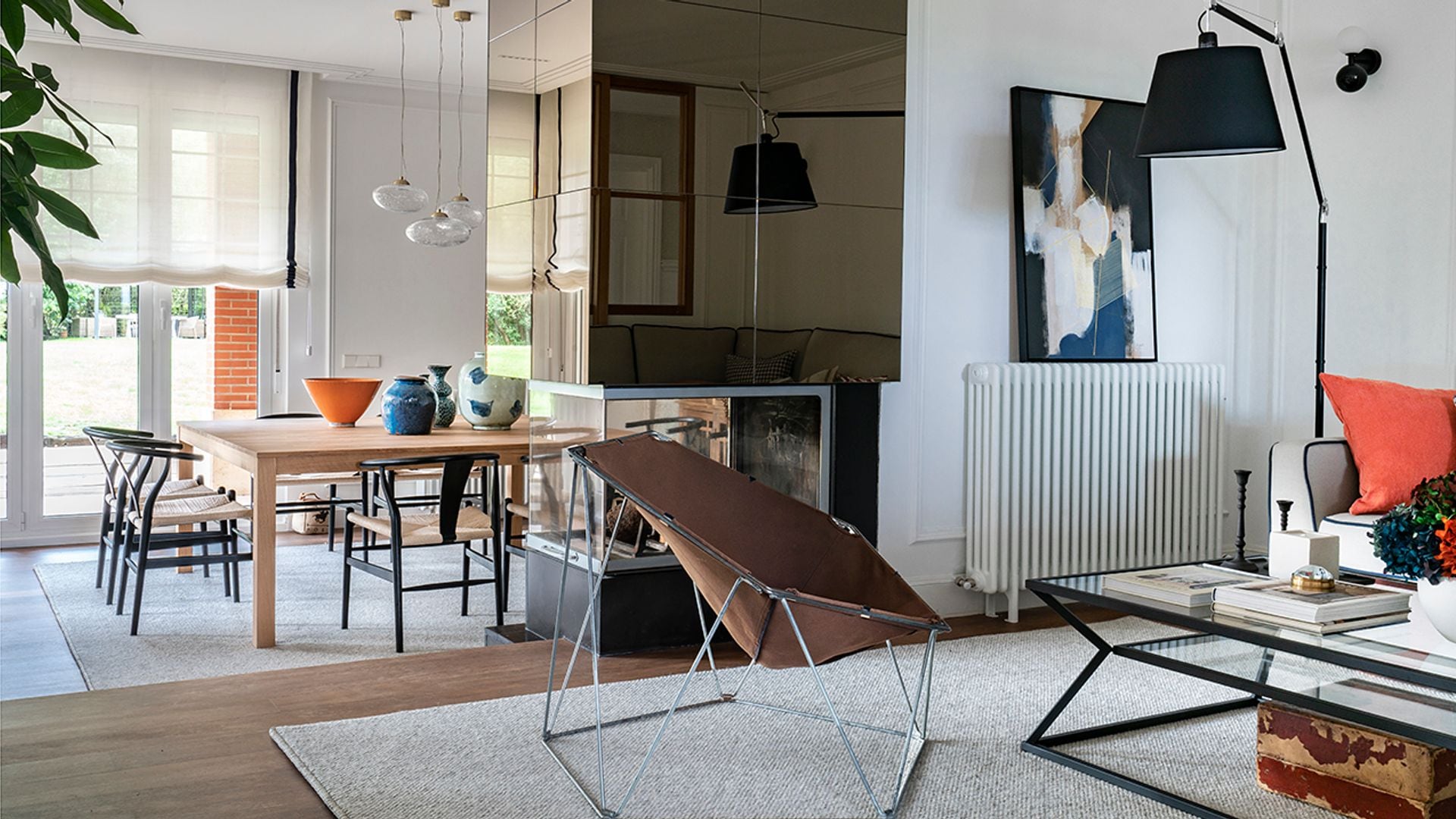 Salón comedor separados por una chimenea de diseño, radiador blanco con cuadro encima, alfombra, lámpara de pie negra, butaca de cuero, mesa de centro de cristal