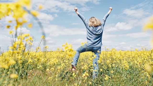 Mujer feliz estirando los brazos hacia arriba en primavera en un campo repleto de pequeñas flores amarillos con el azul cielo detrás