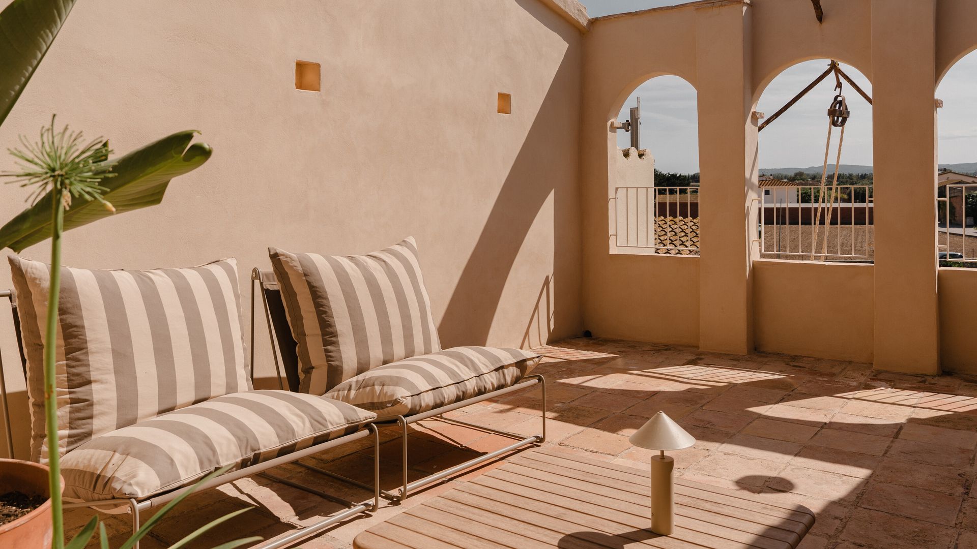 Terraza con suelo de barro, sofás con cojines de rayas, mesa de centro de madera, lámpara portátil 