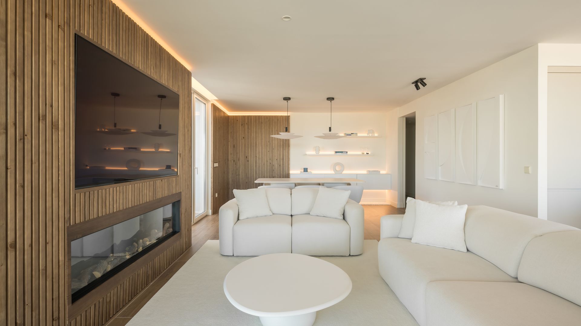 Salón con chimenea moderna, sofás blancos, alfombra blanca, mesa de centro blanca, estructura de madera que integra la televisión y la chimenea, luz perimetral 