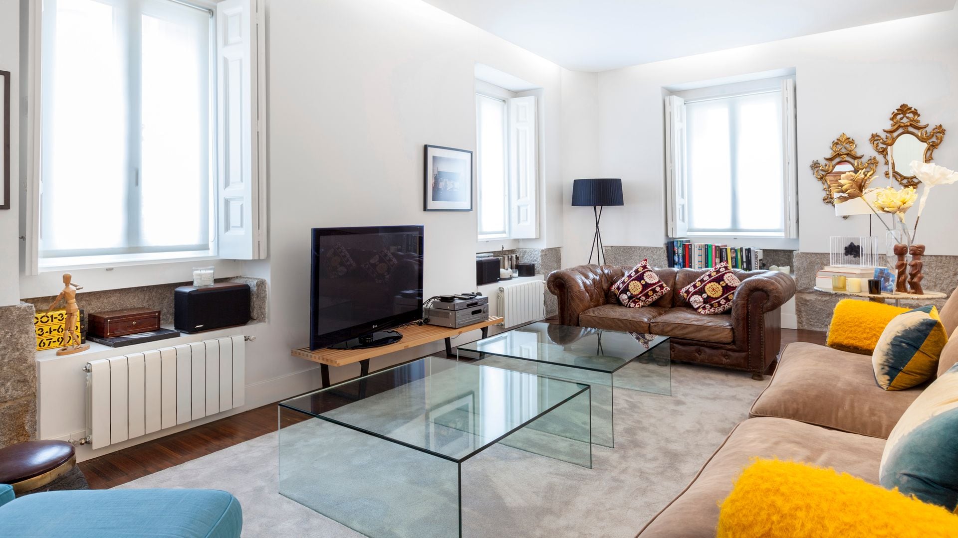 Salón con sofá Chester de cuero marrón, mesas de centro de metacrilato transparente, alfombra, dos radiadores, ventanas con contraventanas de madera blancas, sofá marrón, cojines amarillos, mueble de televisión