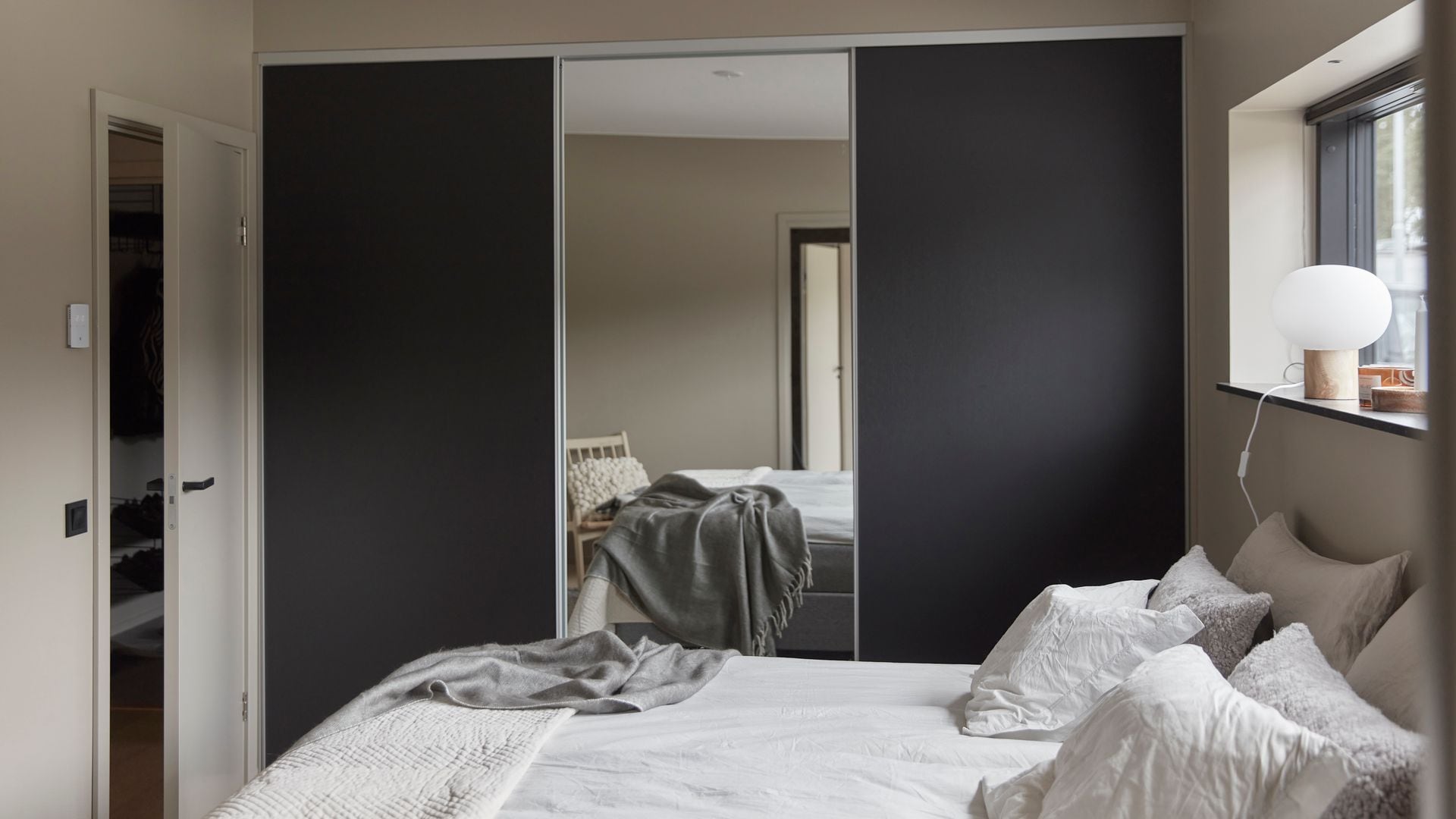 Dormitorio con armario negro con puerta central de espejo, cama sin cabecero, ropa de cama blanca, alféizar ventana con lámpara de sobremesa 