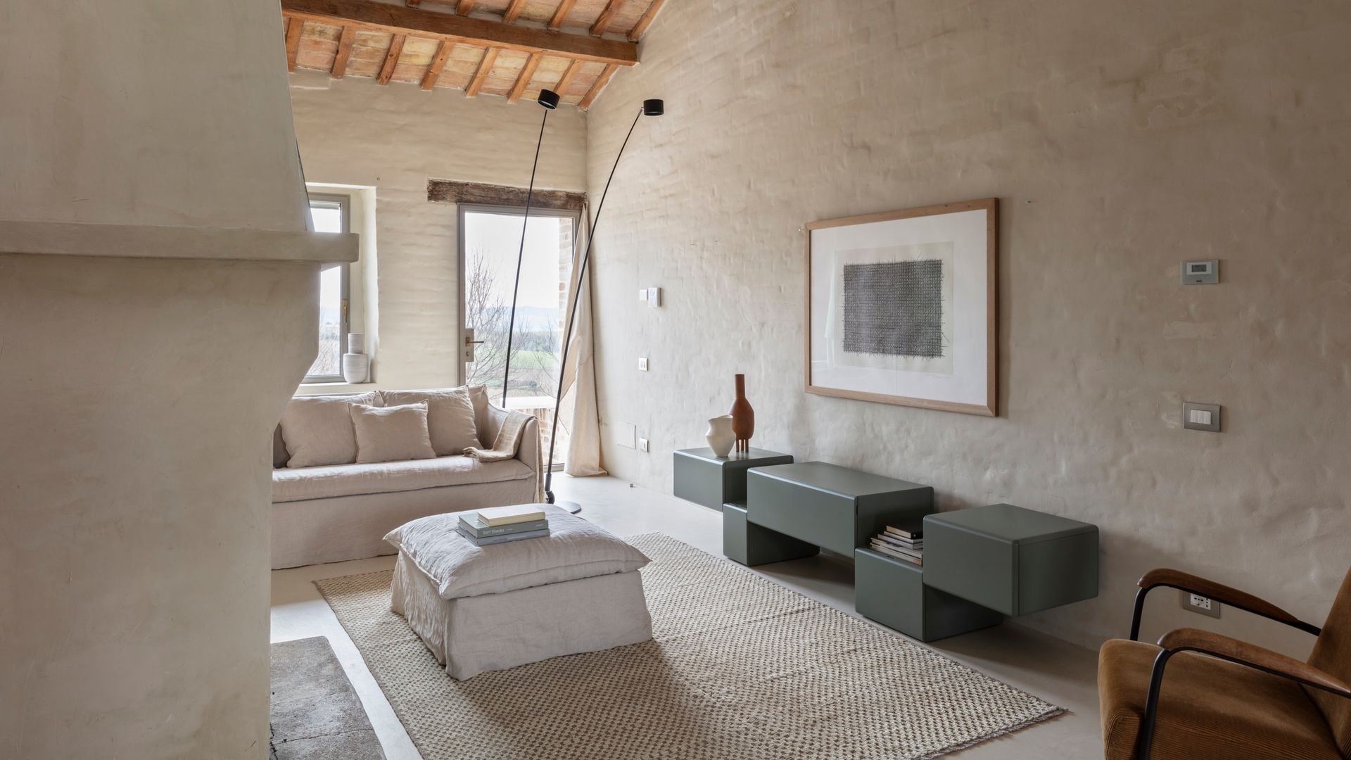 Salón con sofá blanco, mesa de centro tapizada en blanco, vigas de madera en el techo, aparador bajo en gris oscuro, alfombra de fibras vegetales, lámparas de pie negras, cuadro en la pared