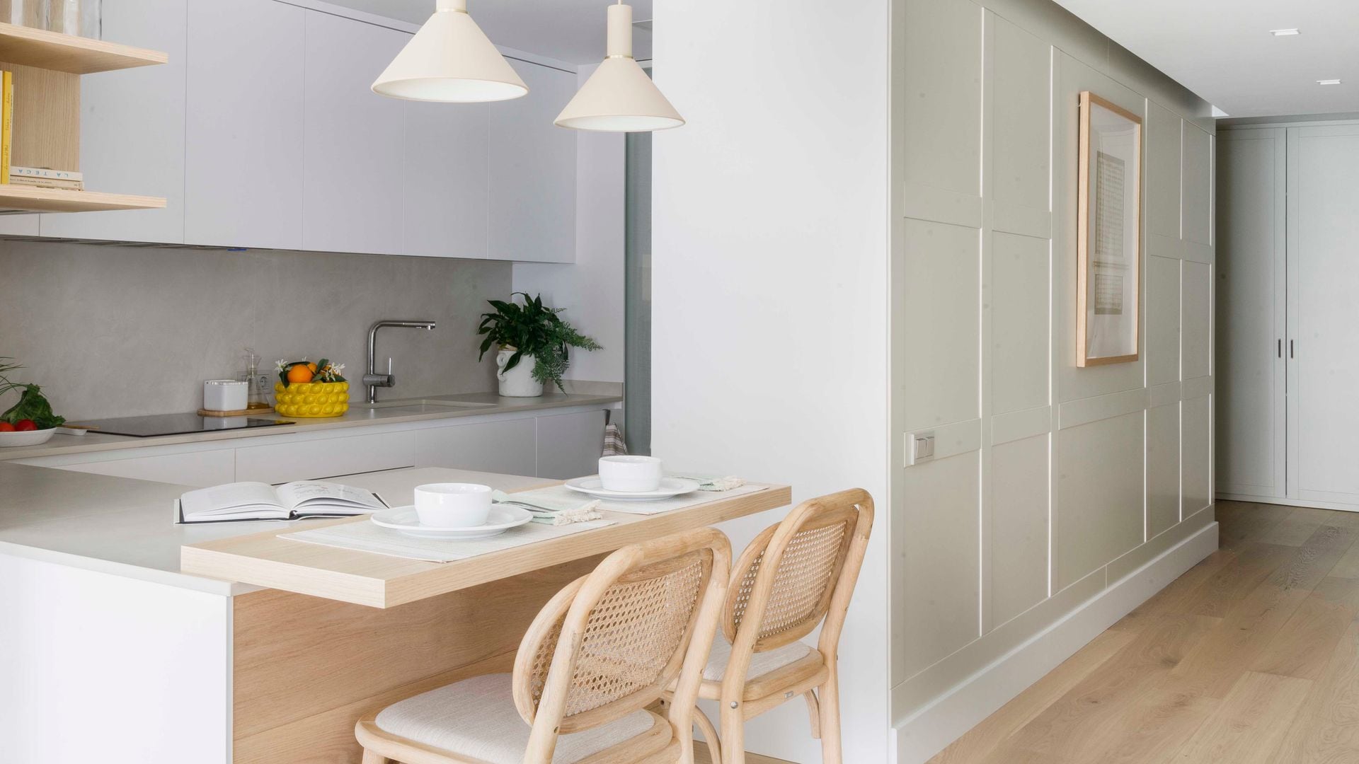 Cocina blanca, abierta con península con barra y dos taburetes con respaldo de rejilla, lámparas de techo, molduras en la pared del recibidor, armario con puertas blancas